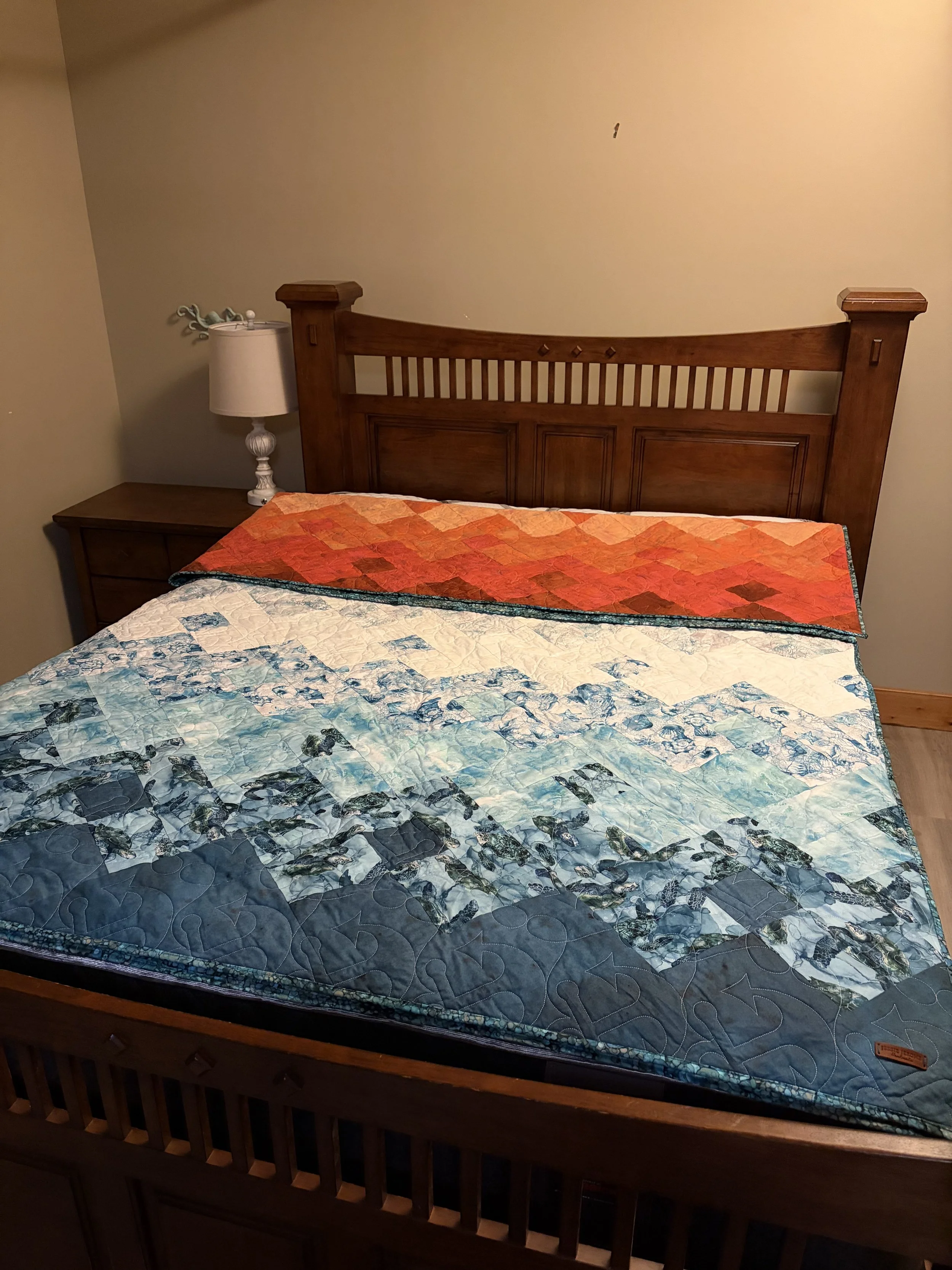 A wooden bed frame with a colorful quilted blanket in shades of orange, red, and blue, and a white and blue patterned quilt at the foot of the bed, beside a nightstand with a white lamp.