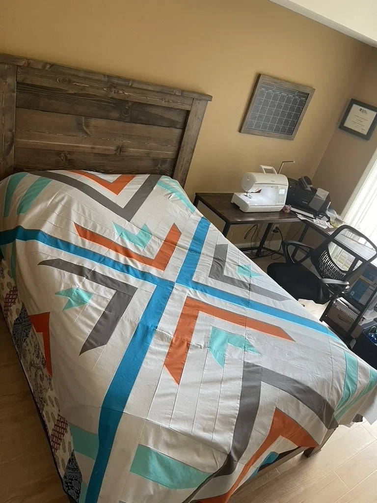 A bedroom with a wooden headboard, featuring a colorful quilt with a geometric pattern in orange, teal, gray, and white. There is a desk with a sewing machine, printer, and other supplies, a black chair, framed certificates, and a grid calendar.