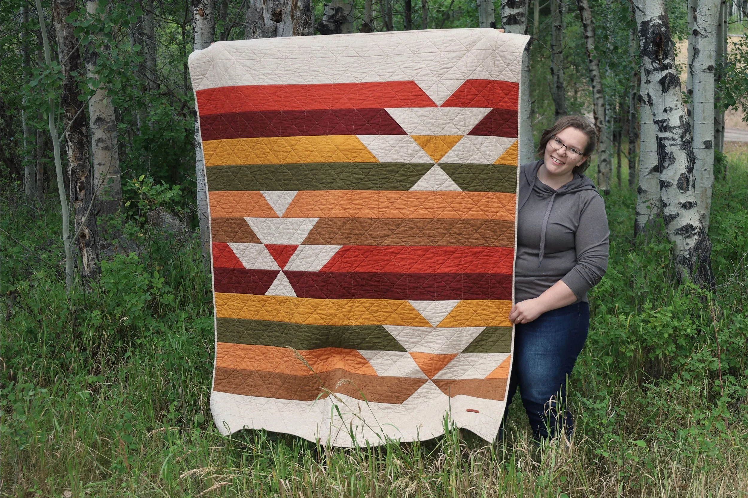 A smiling woman standing outdoors in a forest holding up a colorful quilt with geometric patterns in shades of orange, red, green, white, and beige.