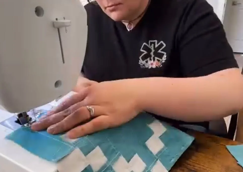 Person sewing a quilt on a sewing machine, wearing a black shirt with medical symbol, rings on fingers, in a room with a white wall and sink.
