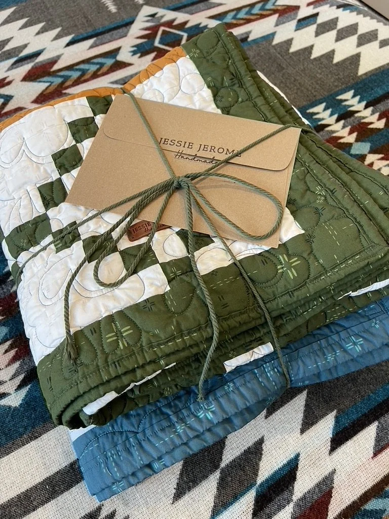 Stack of handmade quilts tied with a green ribbon and a Jesse Jerome handmade branding tag, placed on a colorful geometric patterned rug.