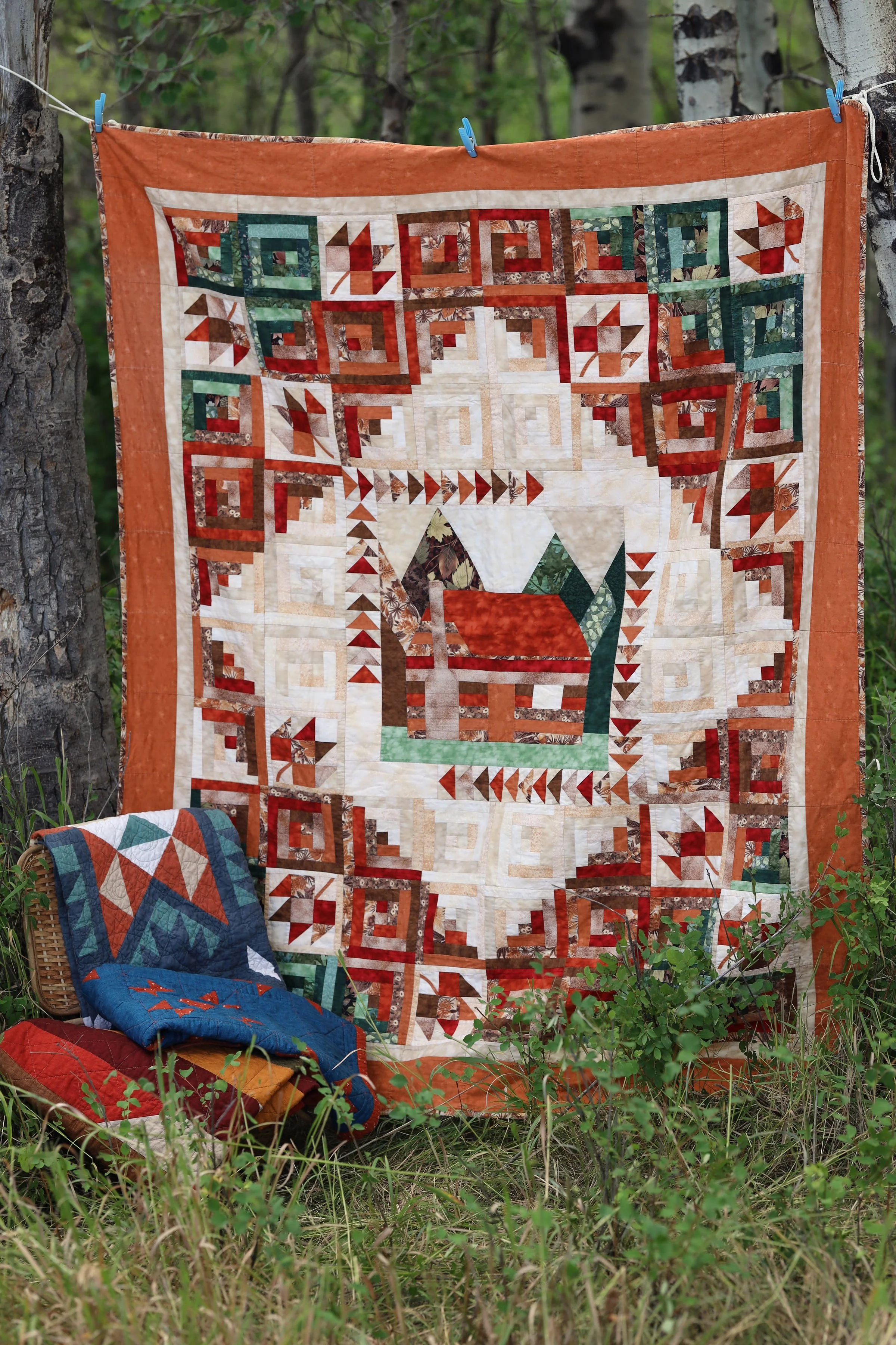 A fabric quilt with intricate geometric and patchwork patterns is hanging between two trees in a forest, with additional quilts draped over a basket and on the ground below. The quilt features a mix of warm colors like orange, red, and brown, with some green and white accents.