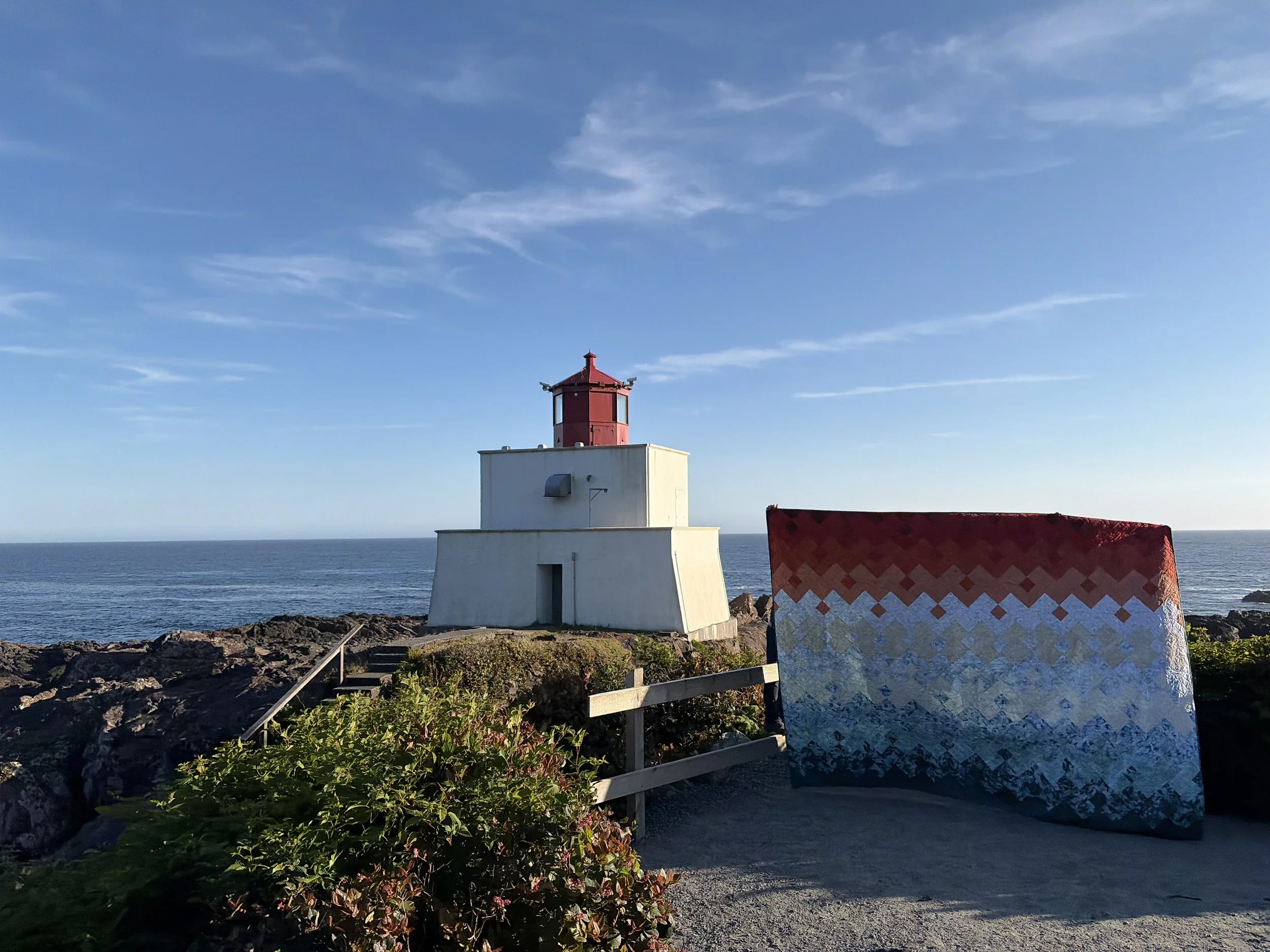 A white lighthouse with a red top standing on rocky shoreline against a blue sky with some clouds, near the ocean, with a colorful blanket or cloth partially visible in the foreground.