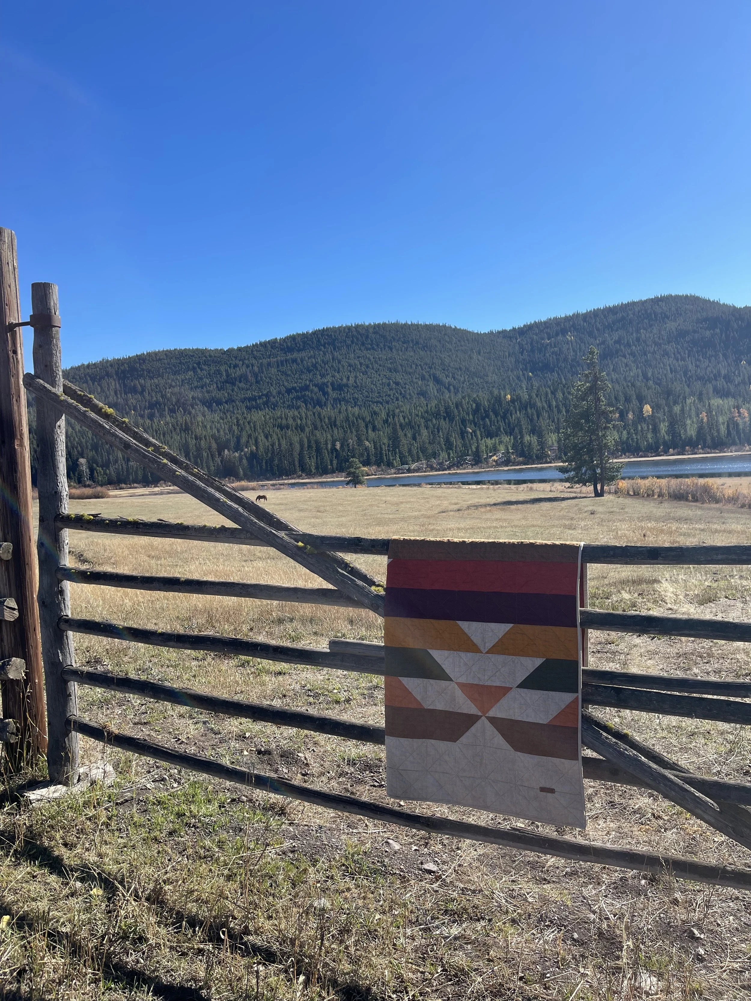 A wooden fence with a colorful quilt hanging from it, overlooking a field, trees, a lake, and mountain hills under a clear blue sky.