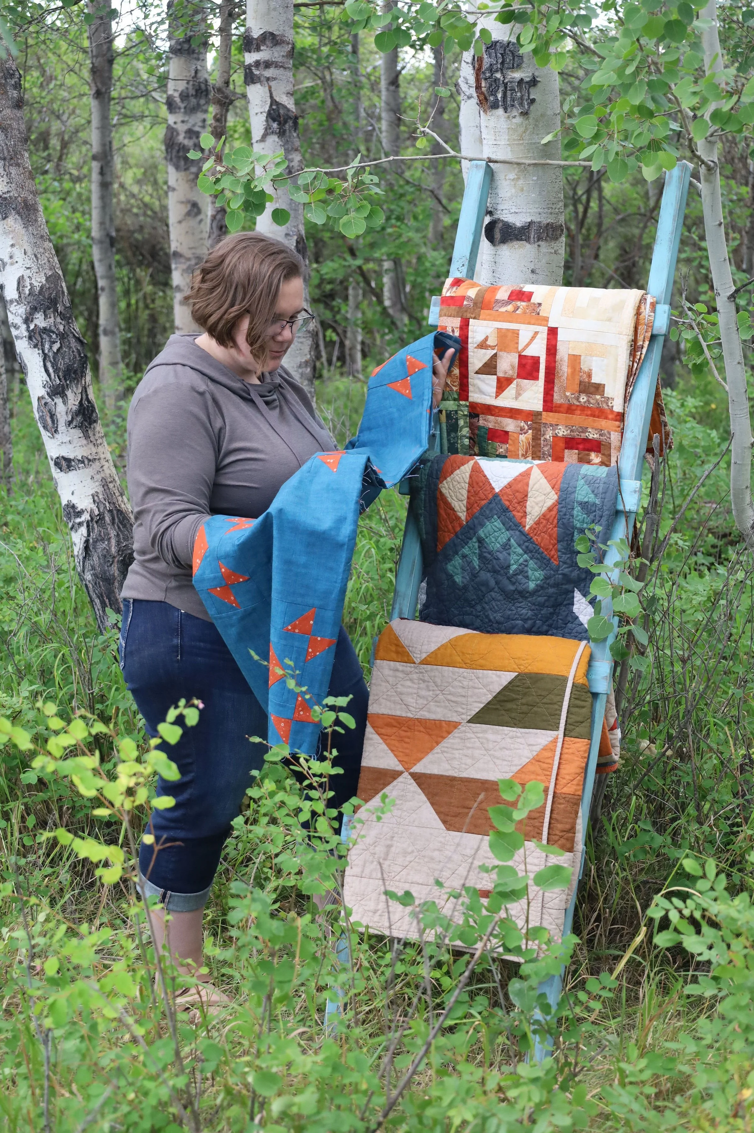 A person with short brown hair, glasses, wearing a gray hoodie and rolled-up jeans, is in a forested area with green foliage. They are holding and examining a colorful quilt with orange, cream, and teal patterns, hanging on a blue wooden ladder leaning against a tree. Another quilt with similar colors is draped over the ladder.