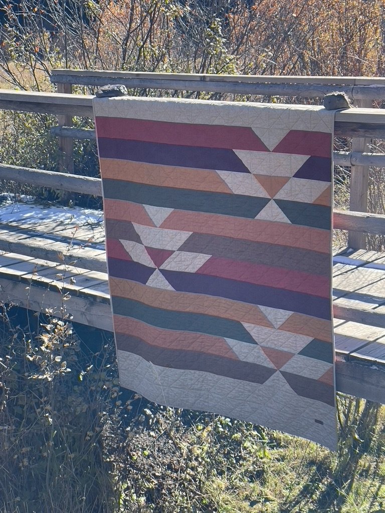 A geometric quilt with a pattern of triangles and parallelograms in shades of red, gray, white, and coral, hanging over a wooden railing outdoors in a wooded area with fall foliage.