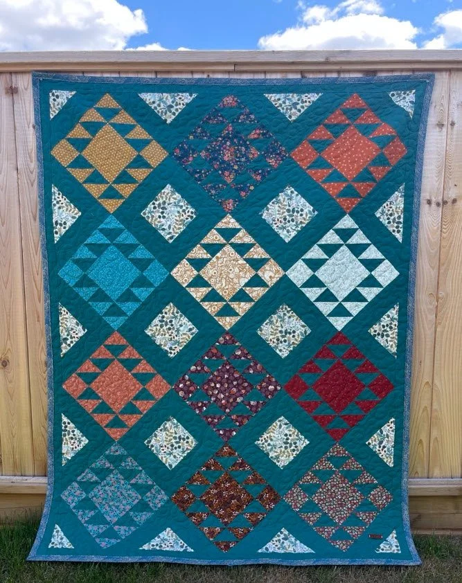 A colorful quilt with a geometric pattern of diamonds and triangles, featuring various floral and patterned fabrics, hanging on a wooden wall outdoors.