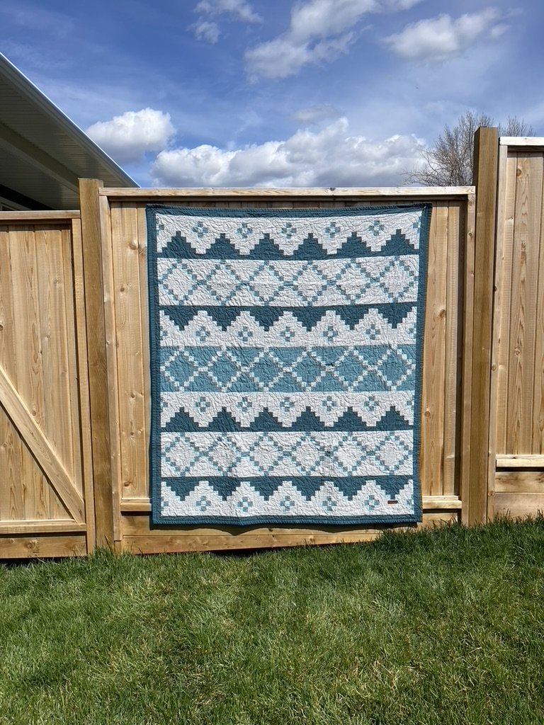 A quilt with geometric patterns in shades of blue, white, and teal hanging on a wooden fence.
