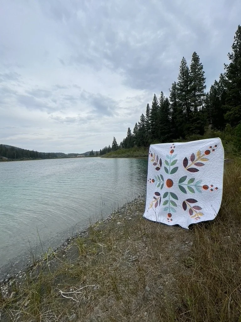 A quilt with a floral pattern is draped over a rock on the grassy bank of a river, with a forested area and cloudy sky in the background.