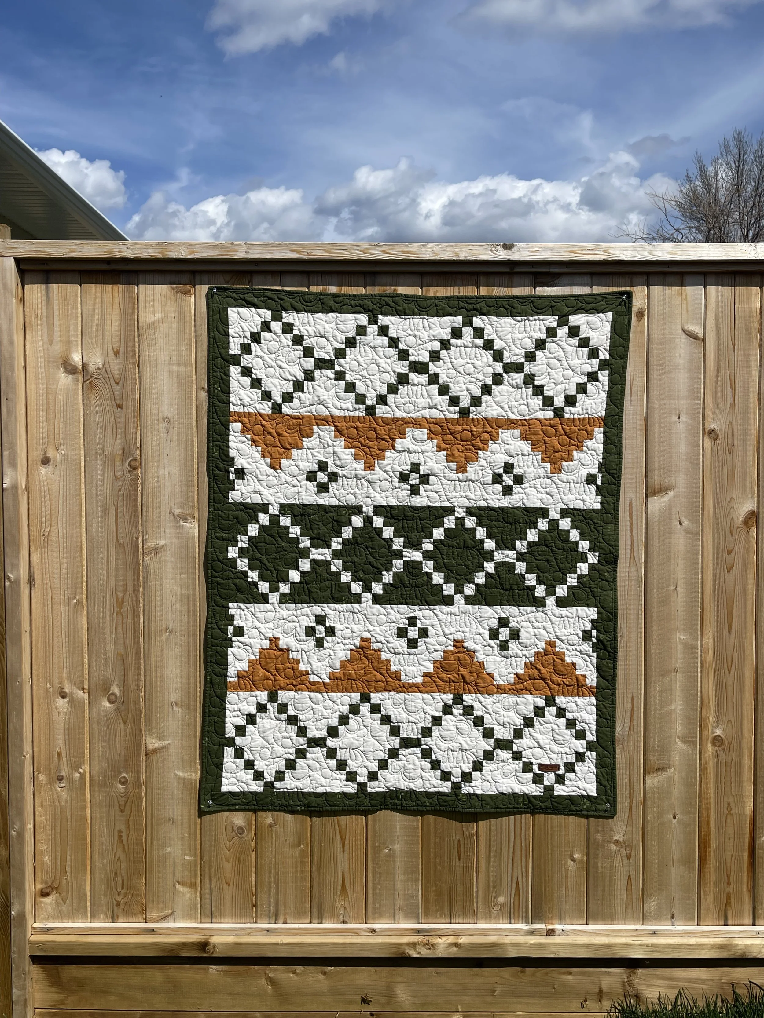 A quilt hanging on a wooden fence outdoors under a partly cloudy sky.