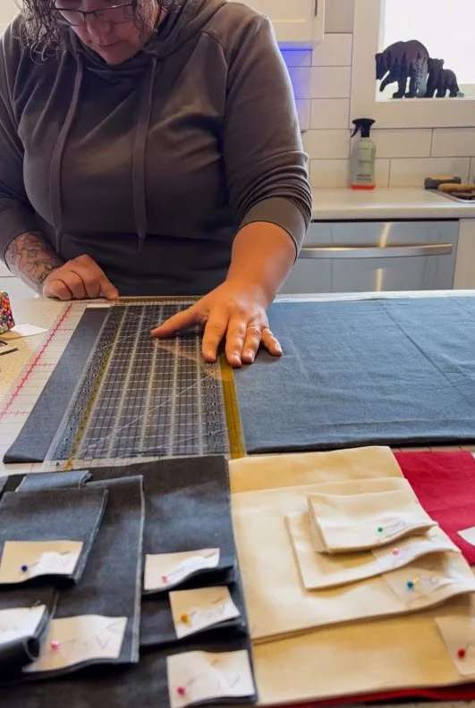 Person using a rotary cutter to cut fabric on a large work table with sewing pin cushions and small fabric pieces in the foreground. Kitchen in the background.
