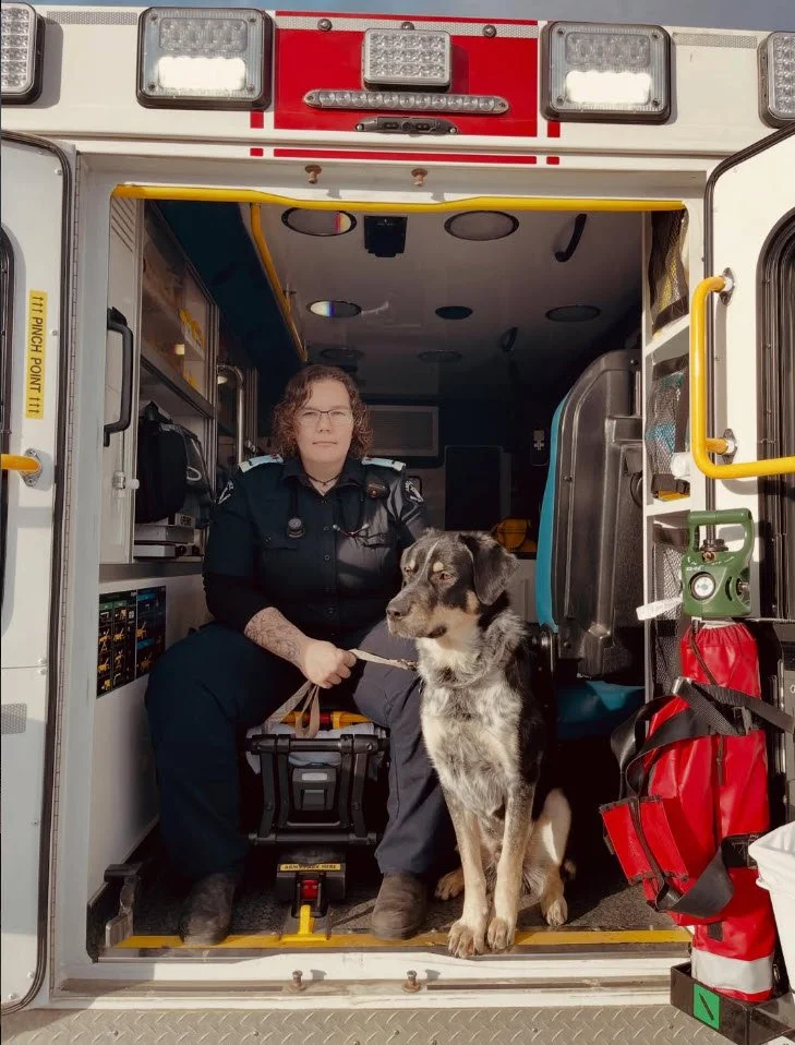 A female paramedic sitting in the back of an ambulance with a black and gray dog on a leash beside her.