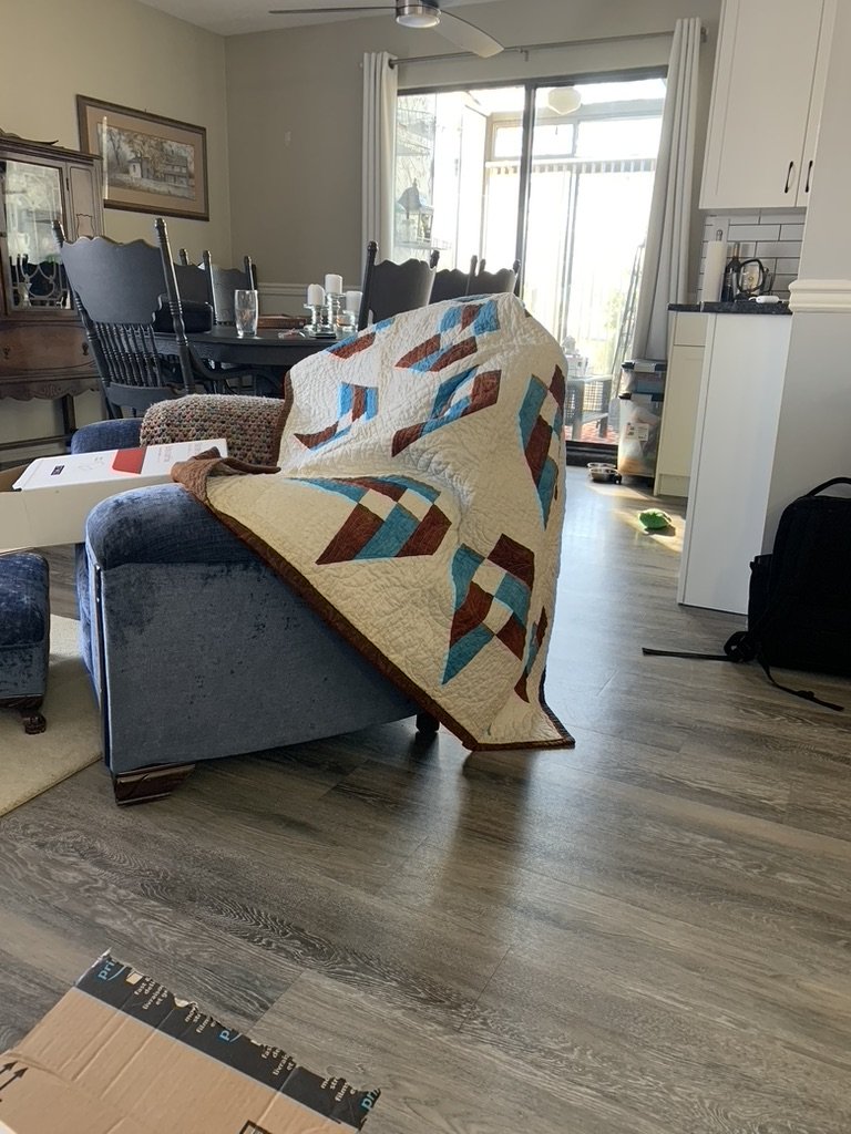 Living room with a blue couch covered by a quilt with geometric patterns in brown, blue, and cream. A dining area with a dark wooden table and chairs, a china cabinet, and a window with white curtains. Kitchen area in the background.