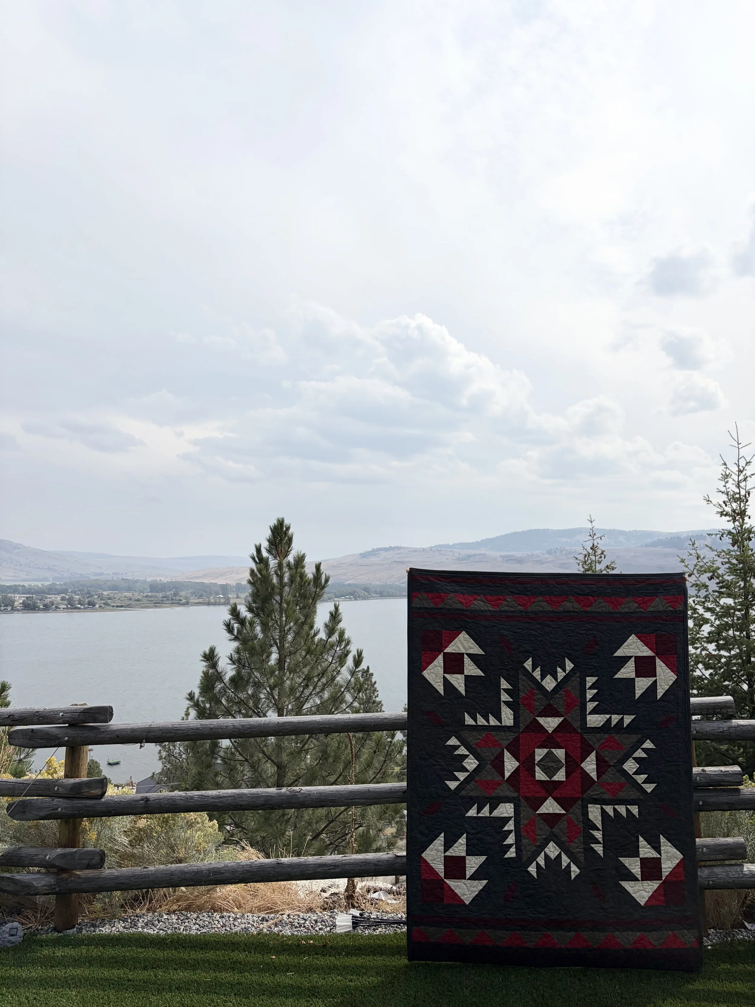 Outdoor scene showing a quilt with a geometric pattern hanging on a wooden fence, overlooking a lake with mountains in the distance under a cloudy sky.