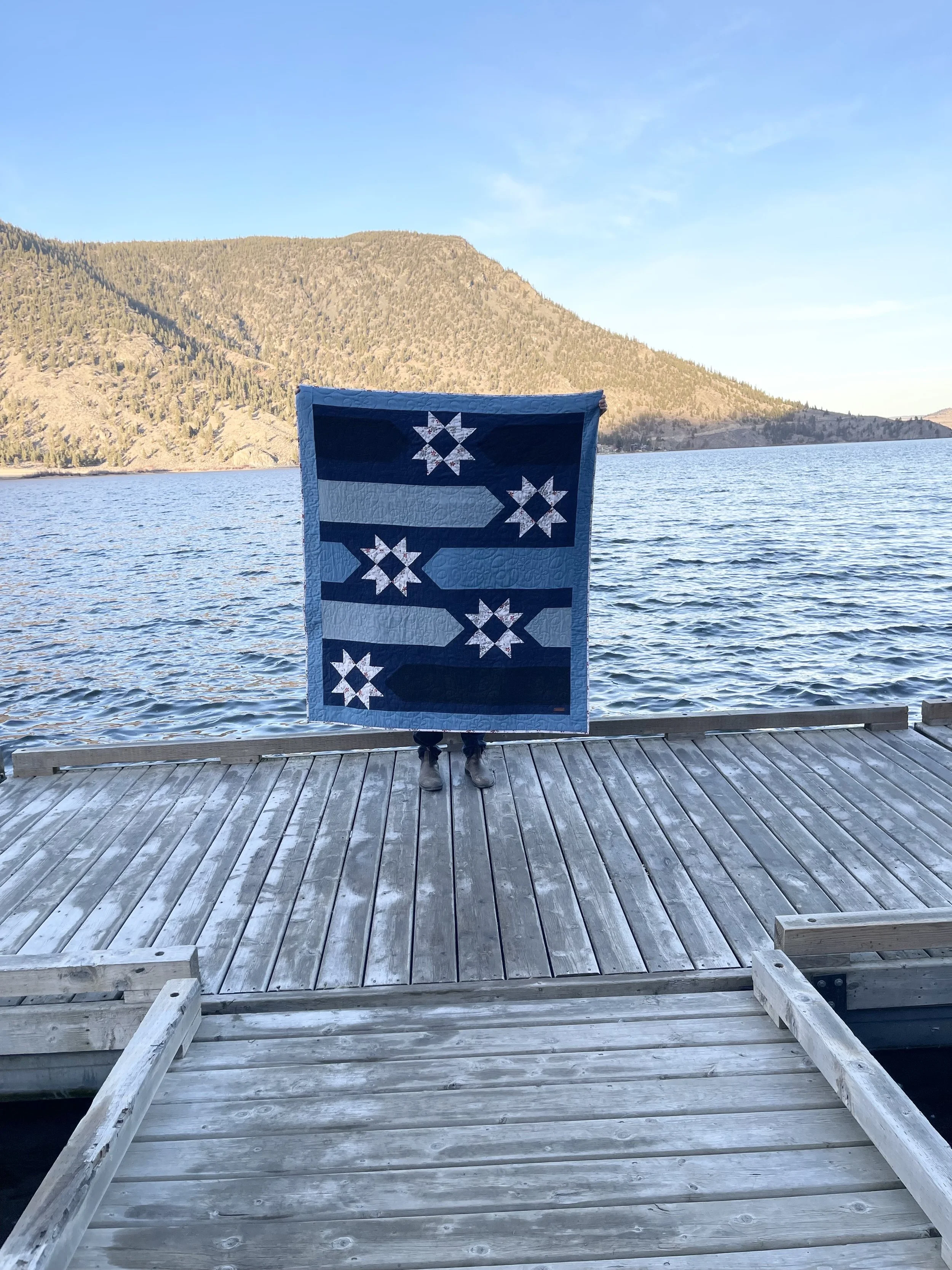 Person holding a quilt with a blue background and star patterns, standing on a wooden dock by a lake with mountains in the background.
