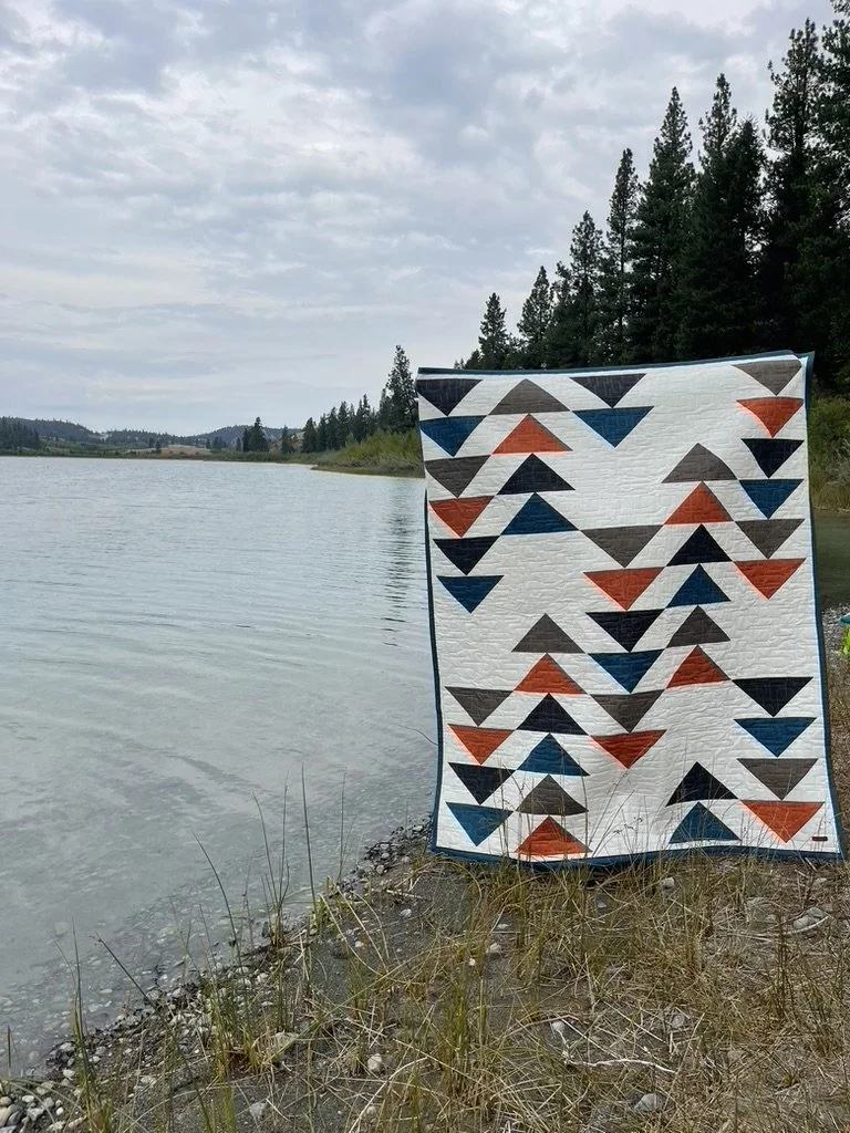 A quilt with a geometric pattern of triangles in blue, orange, black, and gray, hanging on a tie at a lakeshore with trees and cloudy sky in the background.