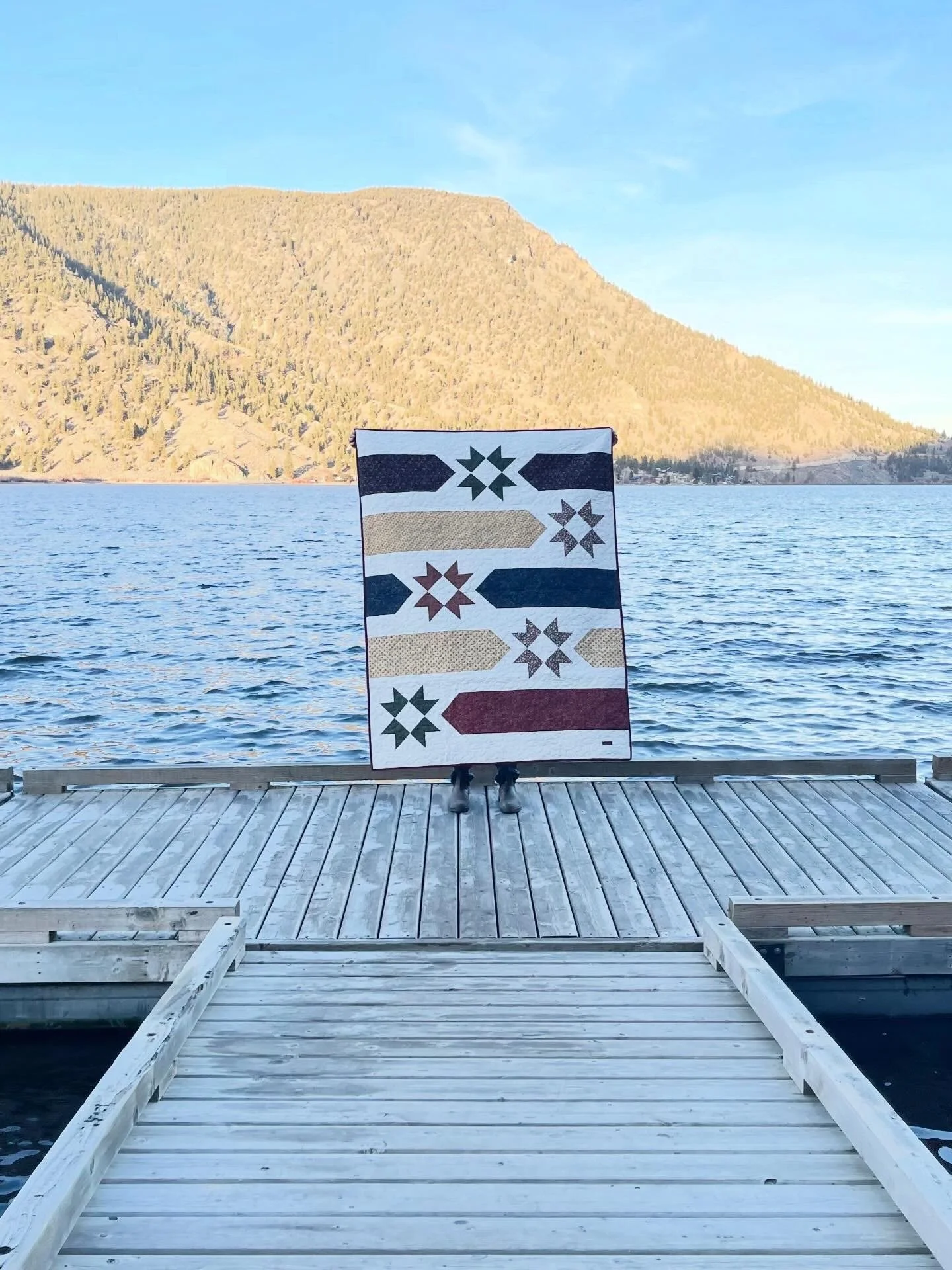 Person holding quilt with arrow and star patterns on a dock over a lake, mountain in background.