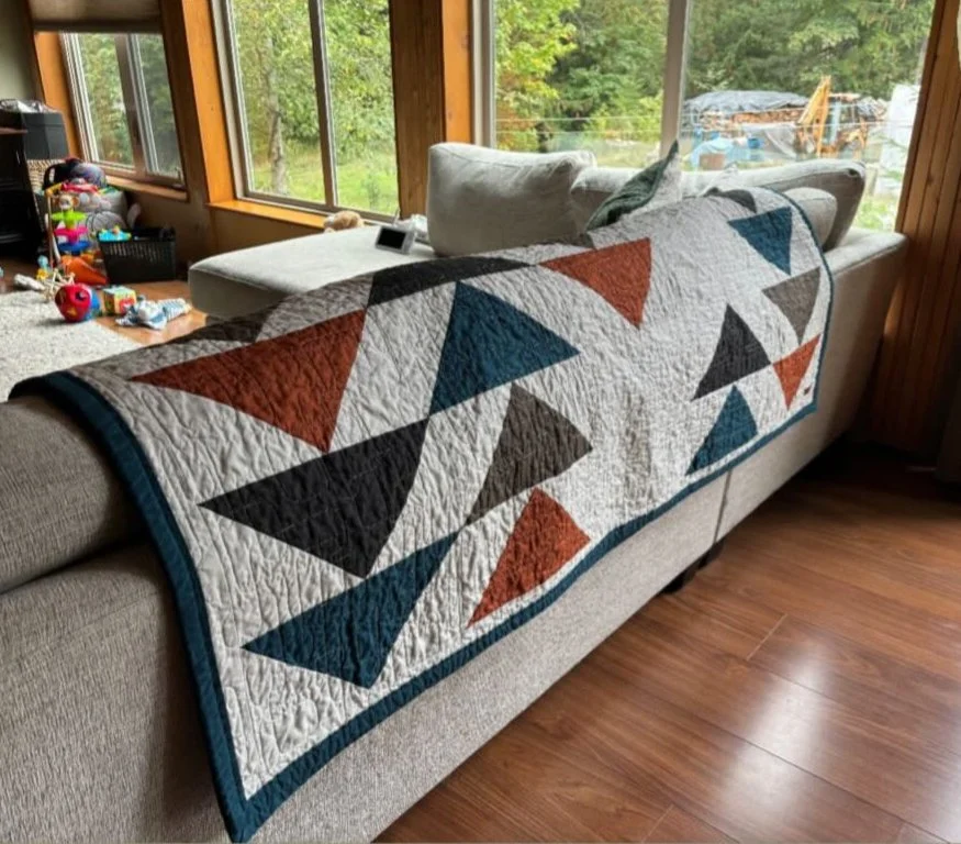 Quilt with geometric triangles in shades of blue, red, brown, and black on the back of a beige couch in a sunlit room with large windows.
