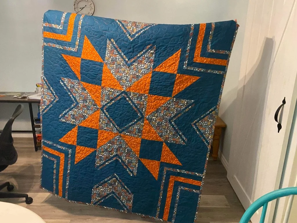 A colorful quilt featuring a star pattern with orange, blue, and floral fabrics, hanging vertically indoors.