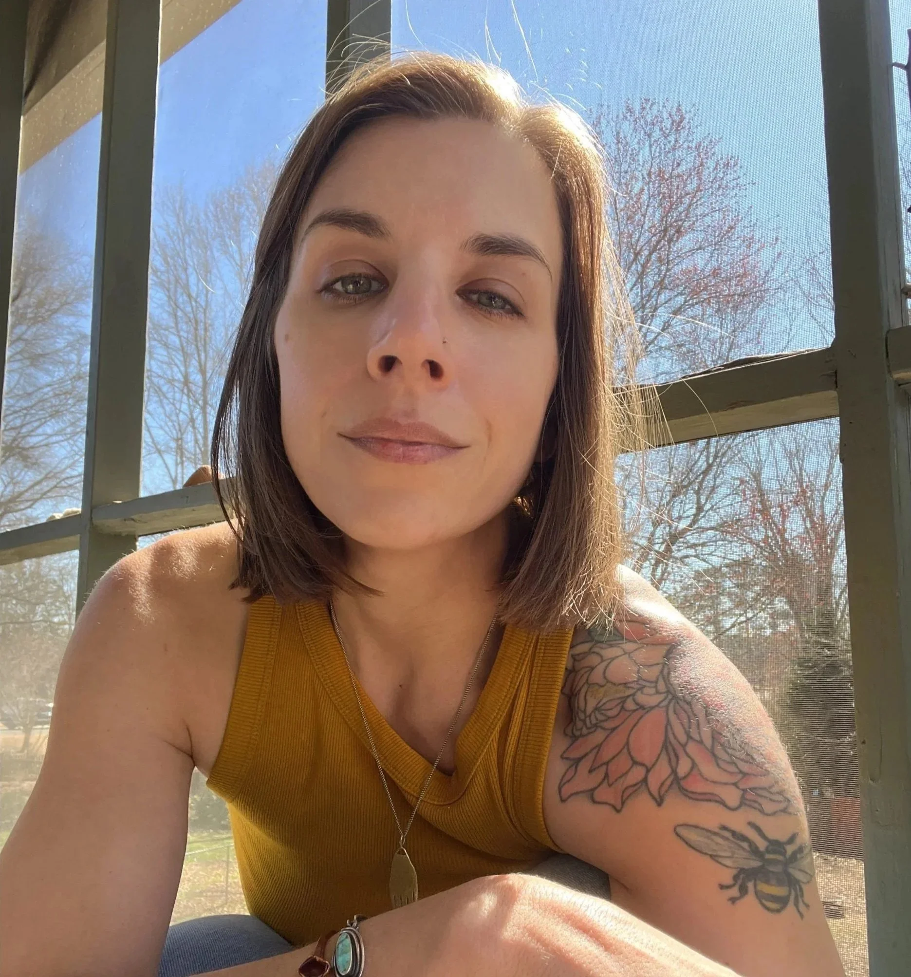 A woman with light brown hair and green eyes taking a selfie outside near a window with a screen. She is wearing a yellow top, a bracelet, and has tattoos on her arm. There are trees with bare branches behind her, and the sky is clear and blue.
