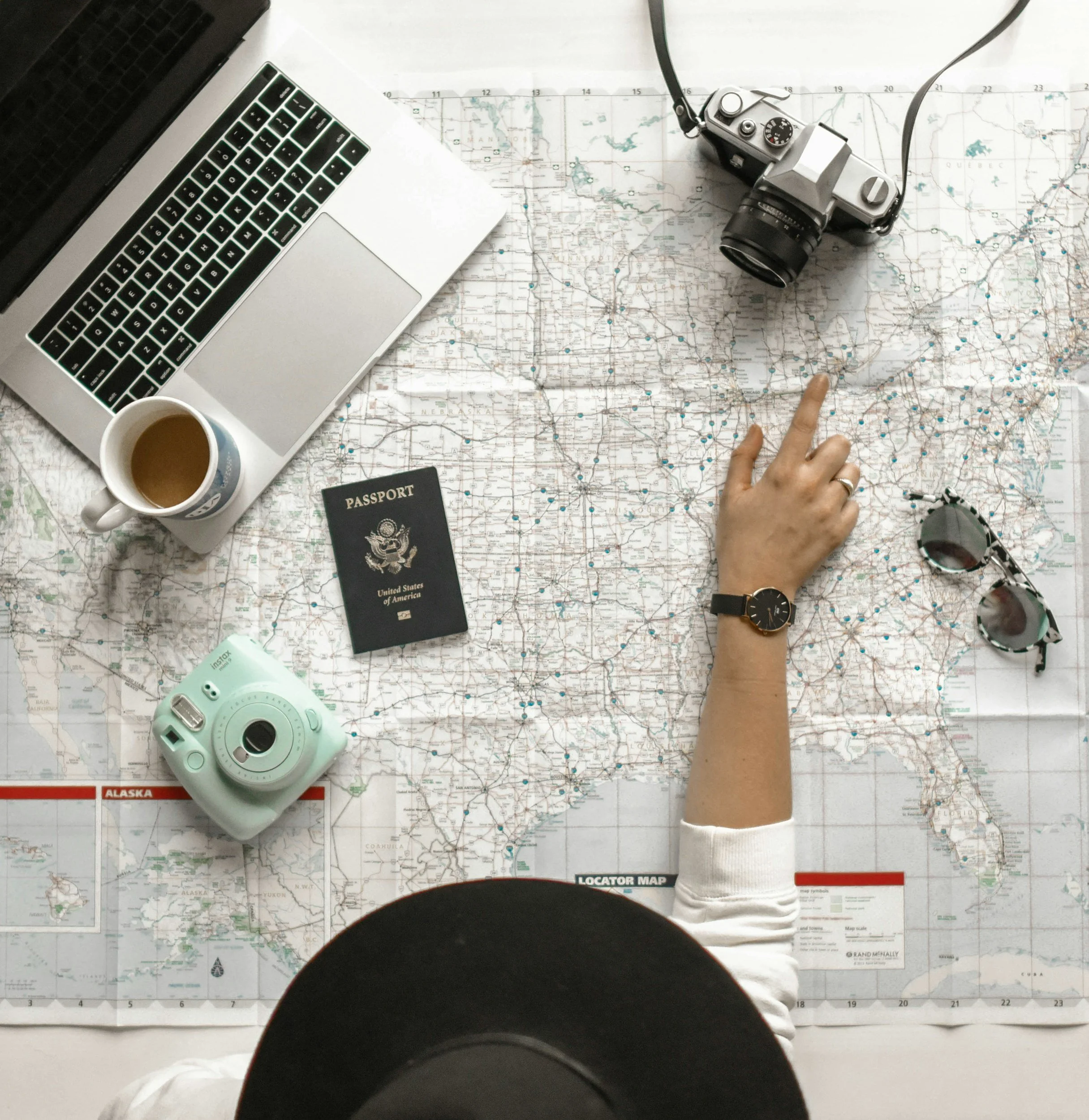 A person pointing at a map of the United States, with a camera, laptop, passport, and a cup of coffee nearby.
