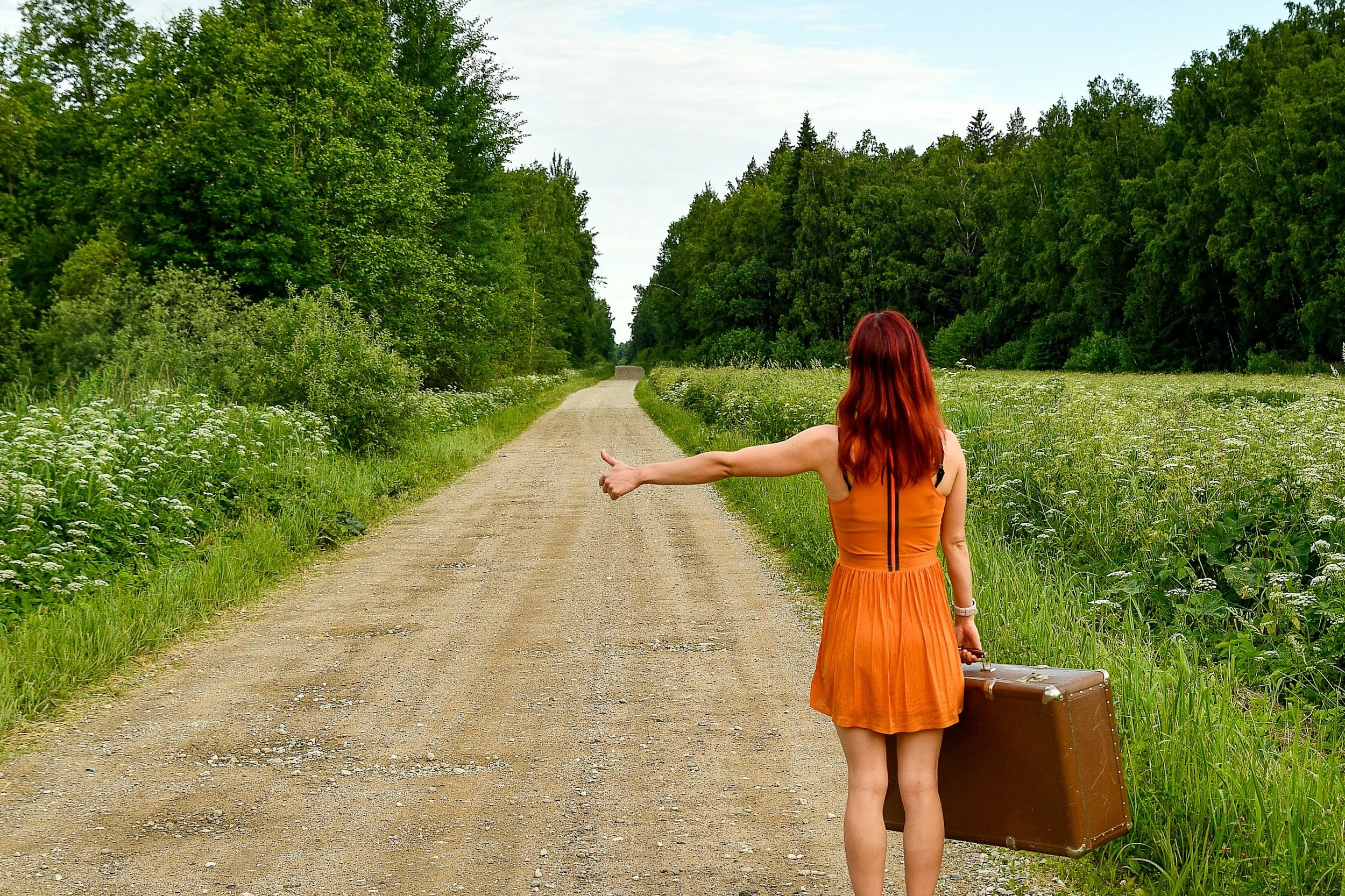 A woman with red hair wearing an orange dress, carrying a brown suitcase, standing on a dirt road in a green, wooded area, with her thumb extended in a hitchhiking gesture.