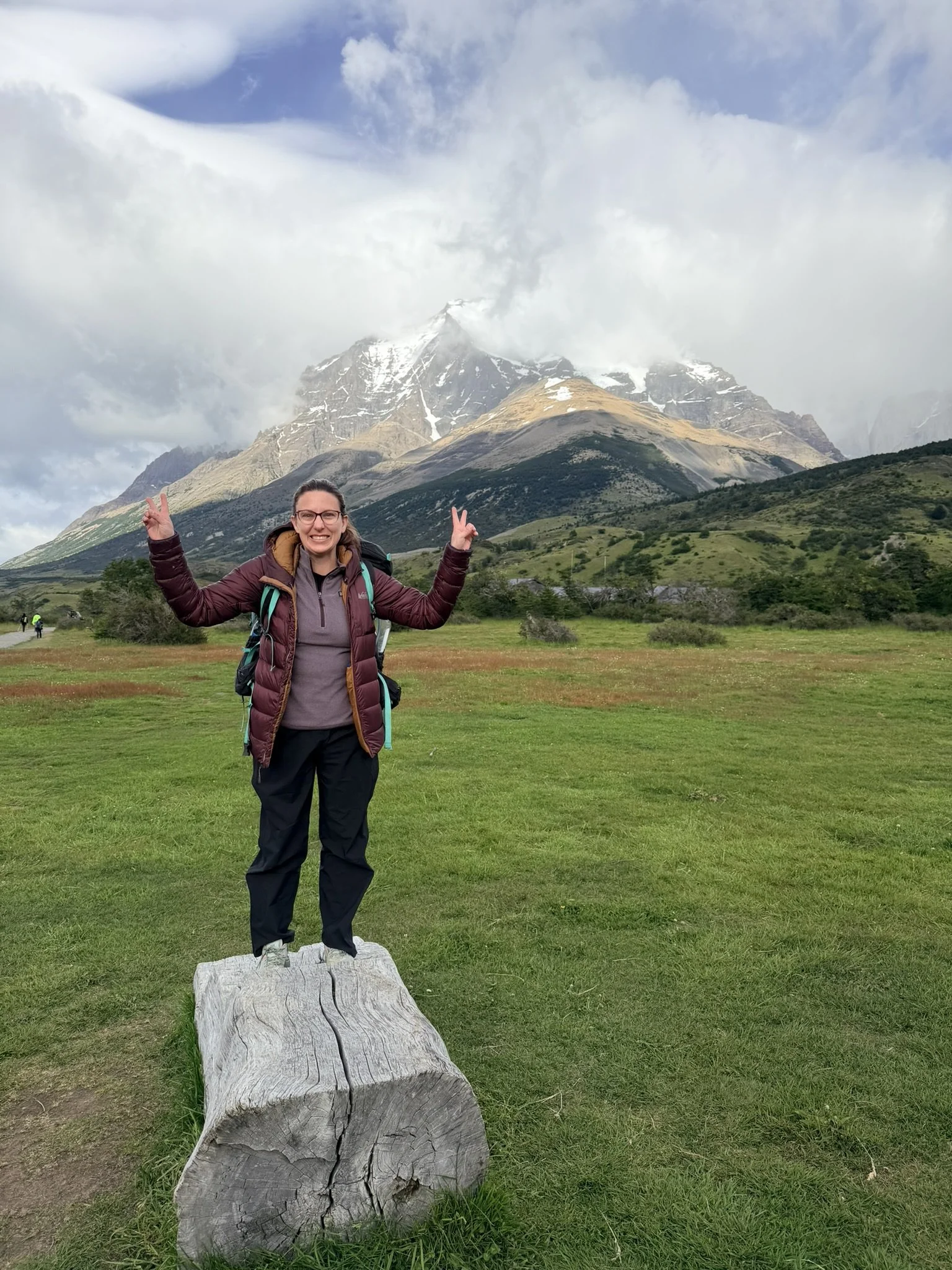A woman standing on a log in front of a mountain range with snow, green hills, cloudy sky, and a person in the distance.