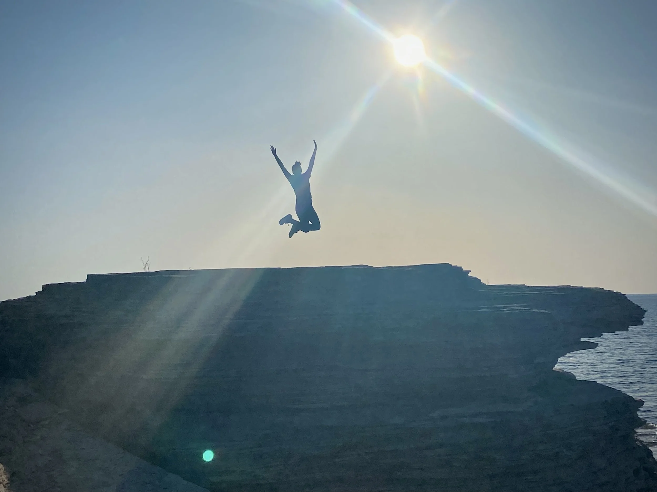 Person jumping in the air on a rocky cliff overlooking a body of water, with the sun shining brightly in the sky and lens flare.