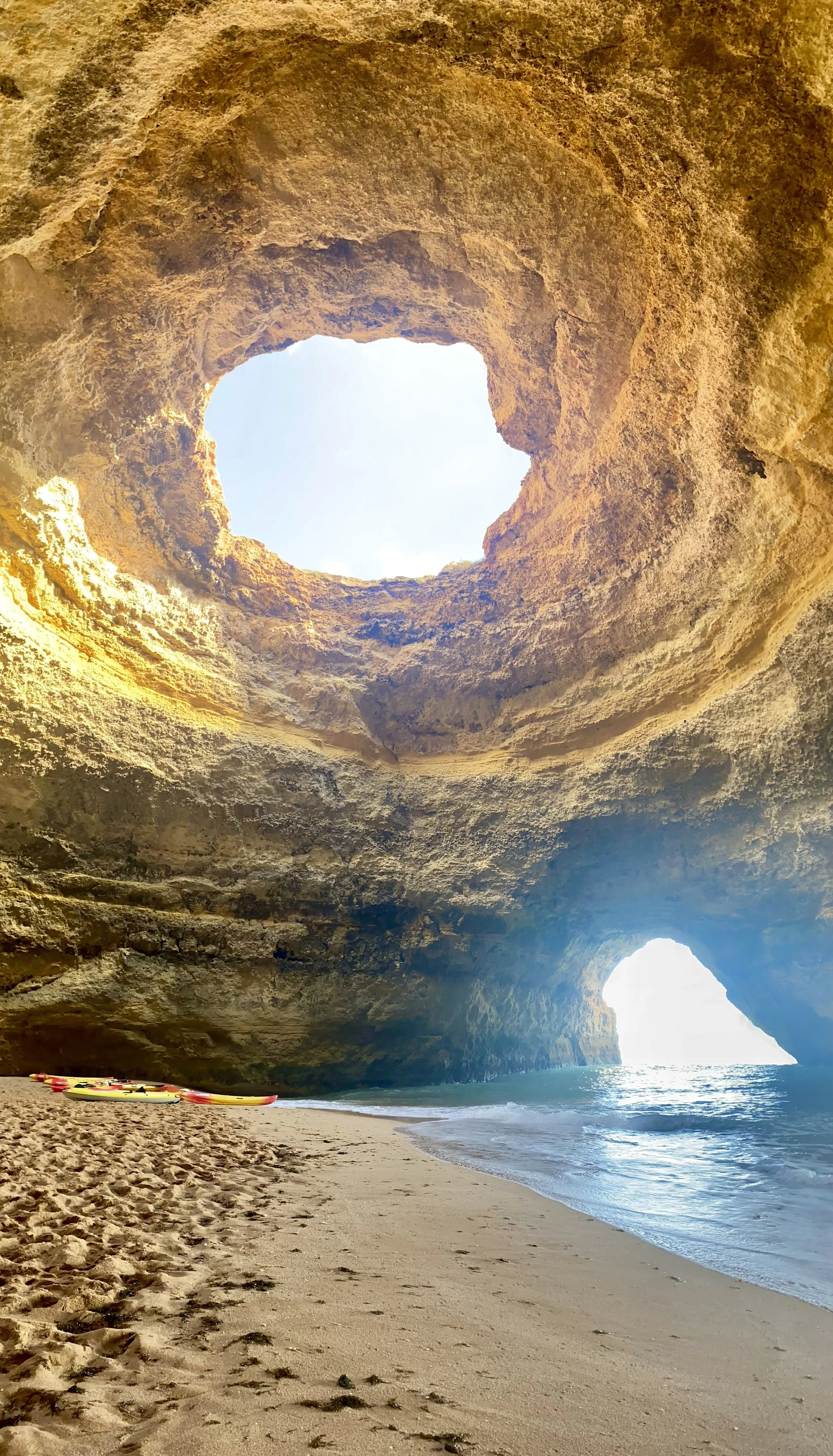 A beach scene with sandy shore, kayaks, and turquoise sea, viewed from inside a large rocky cave with openings showing blue sky.