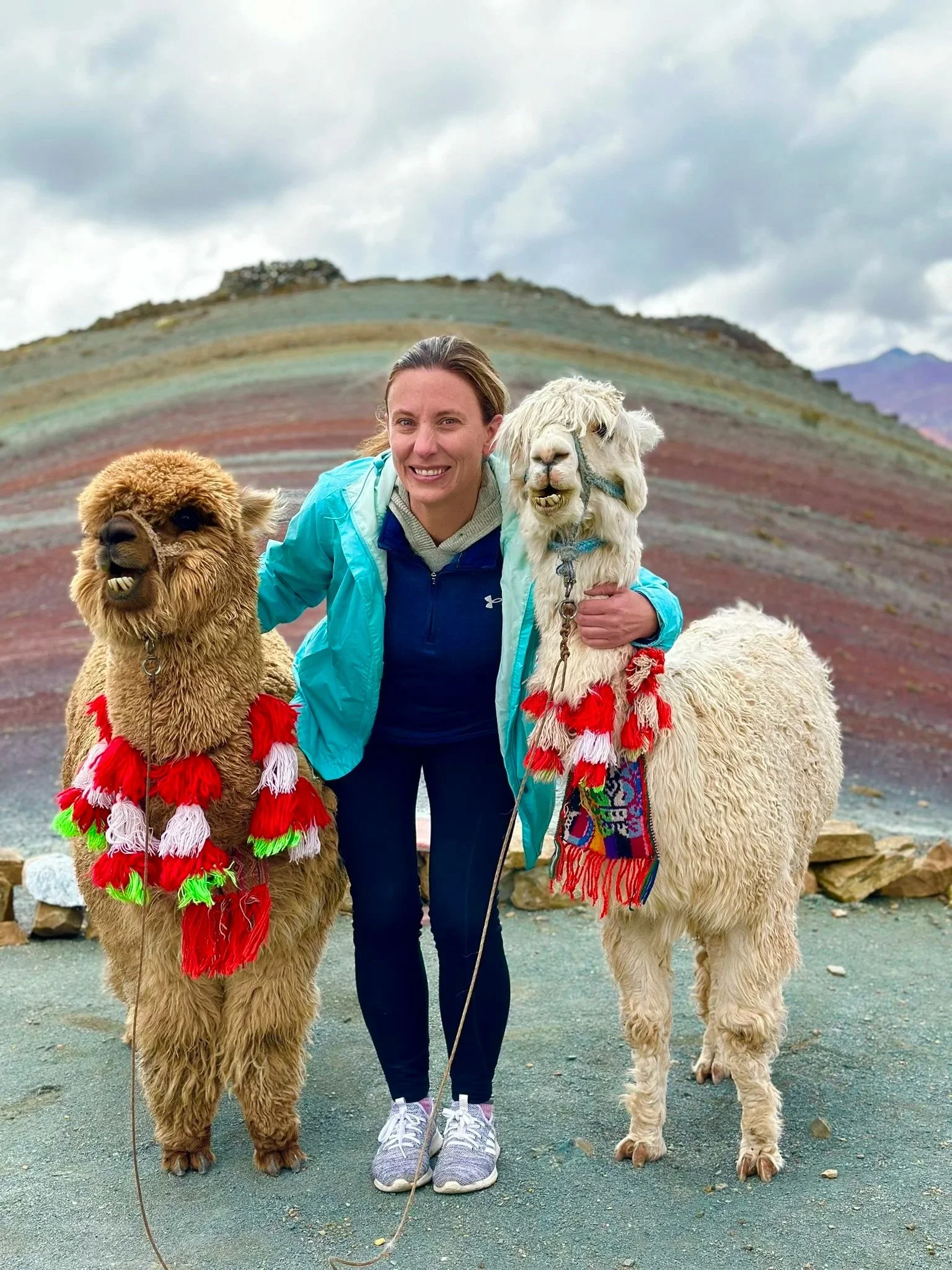 A woman in a turquoise jacket and sneakers with a woman and two llamas, one brown and one white, with colorful scarves around their necks, standing on a mountain trail under cloudy skies.