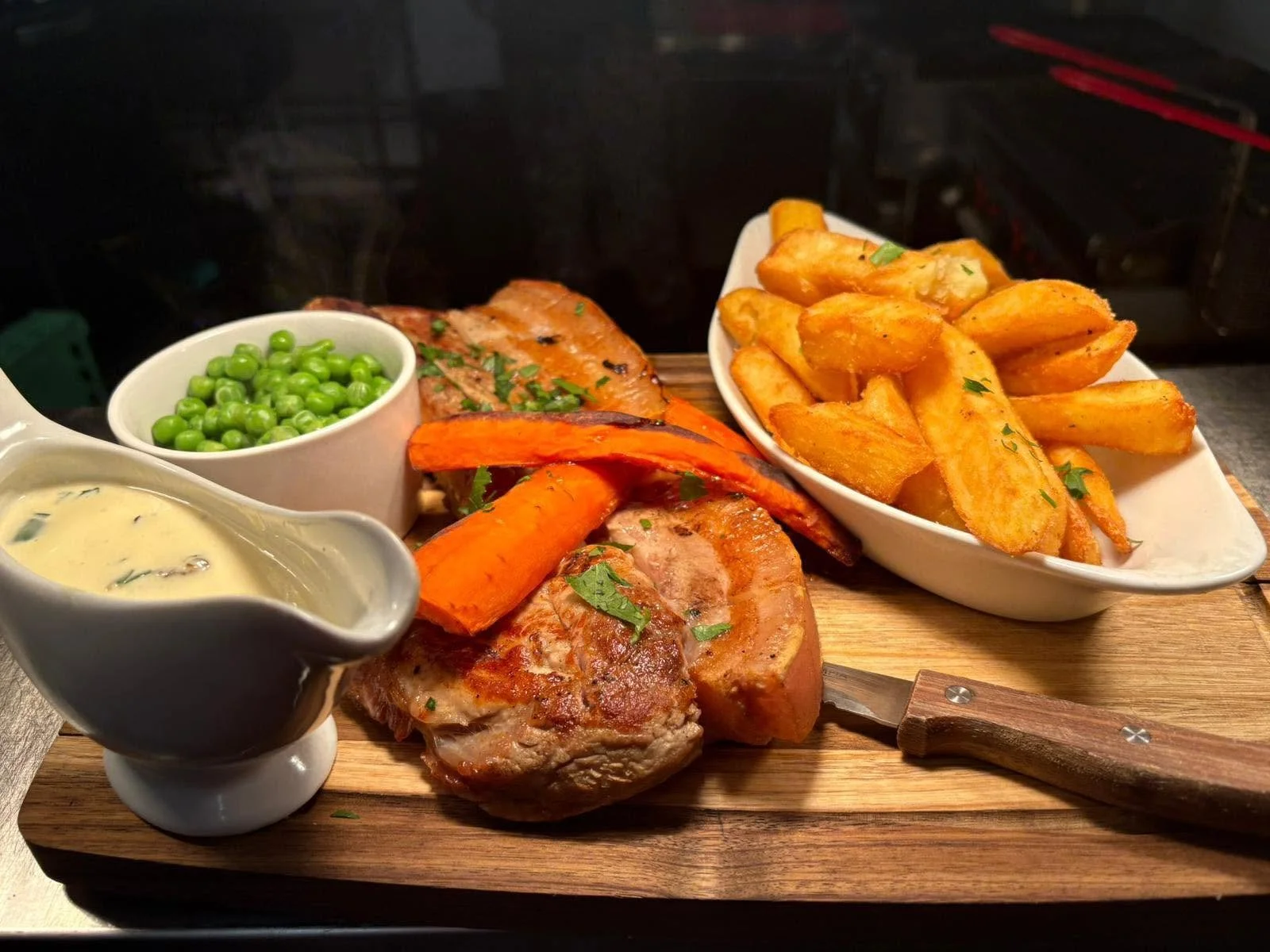 A serving of roasted meat with grilled carrots, green peas, potato wedges, and sauce on a wooden board.