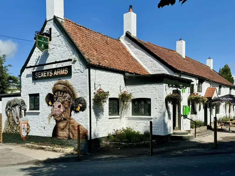 A white building with a red-tiled roof, decorated with murals of animals, including a sheep with ram horns and a boar with tusks, and a sign that reads 'Sexey's Arms'. There are hanging flower baskets, an outdoor seating area, and a green phone box near the entrance.