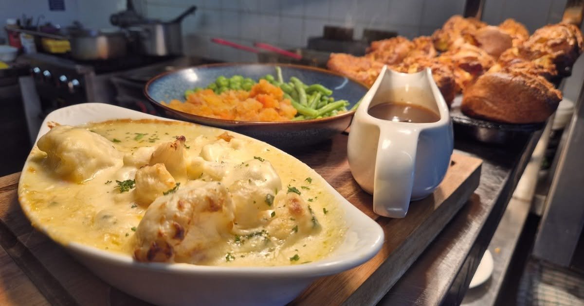 A serving of creamy cauliflower, a bowl of mashed sweet potatoes and green beans, and a jug of gravy with roasted chicken and cornbread in the background.