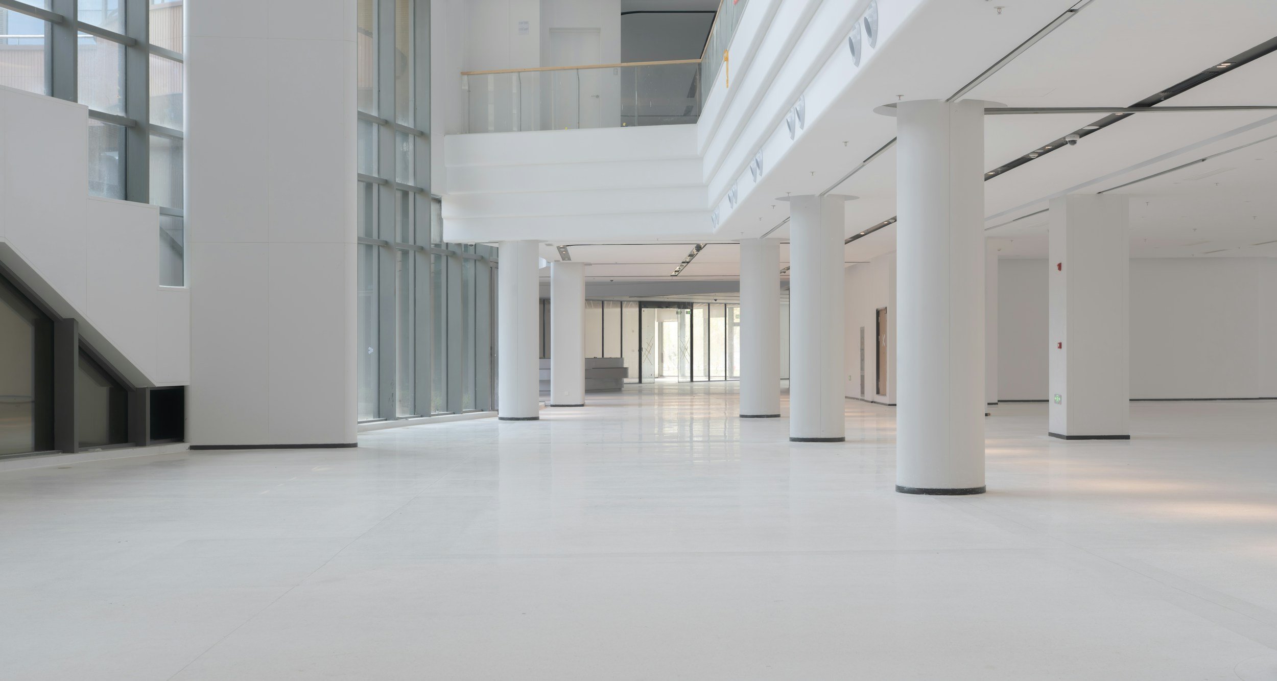 A spacious, modern, and empty interior with white walls, large glass windows, white columns, and a high ceiling, possibly in a commercial or office building.