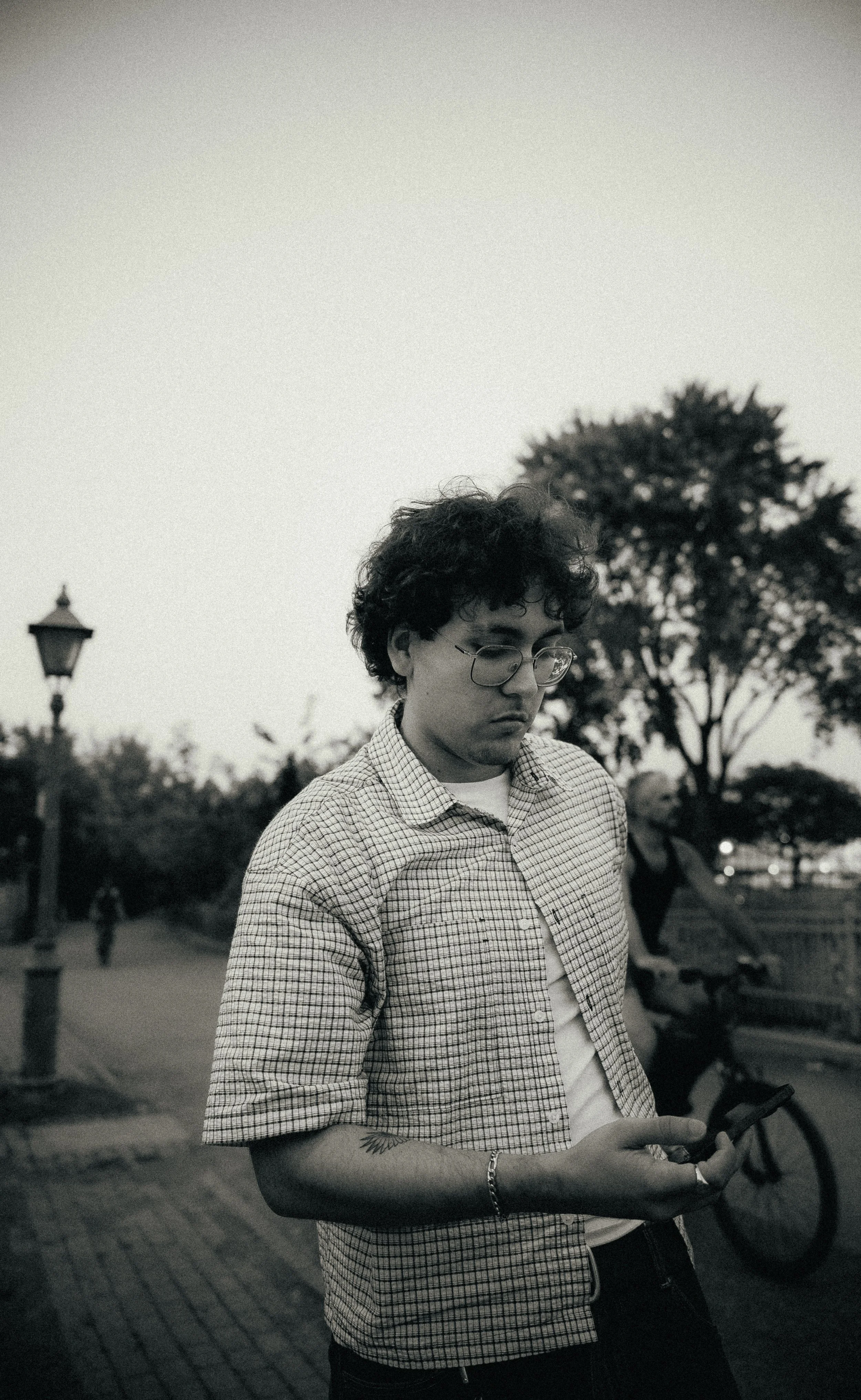 Un jeune homme portant des lunettes, une chemise à carreaux, regarde son téléphone dans un parc ou une zone publique en soirée, avec des arbres, un lampadaire et des autres personnes en arrière-plan.
