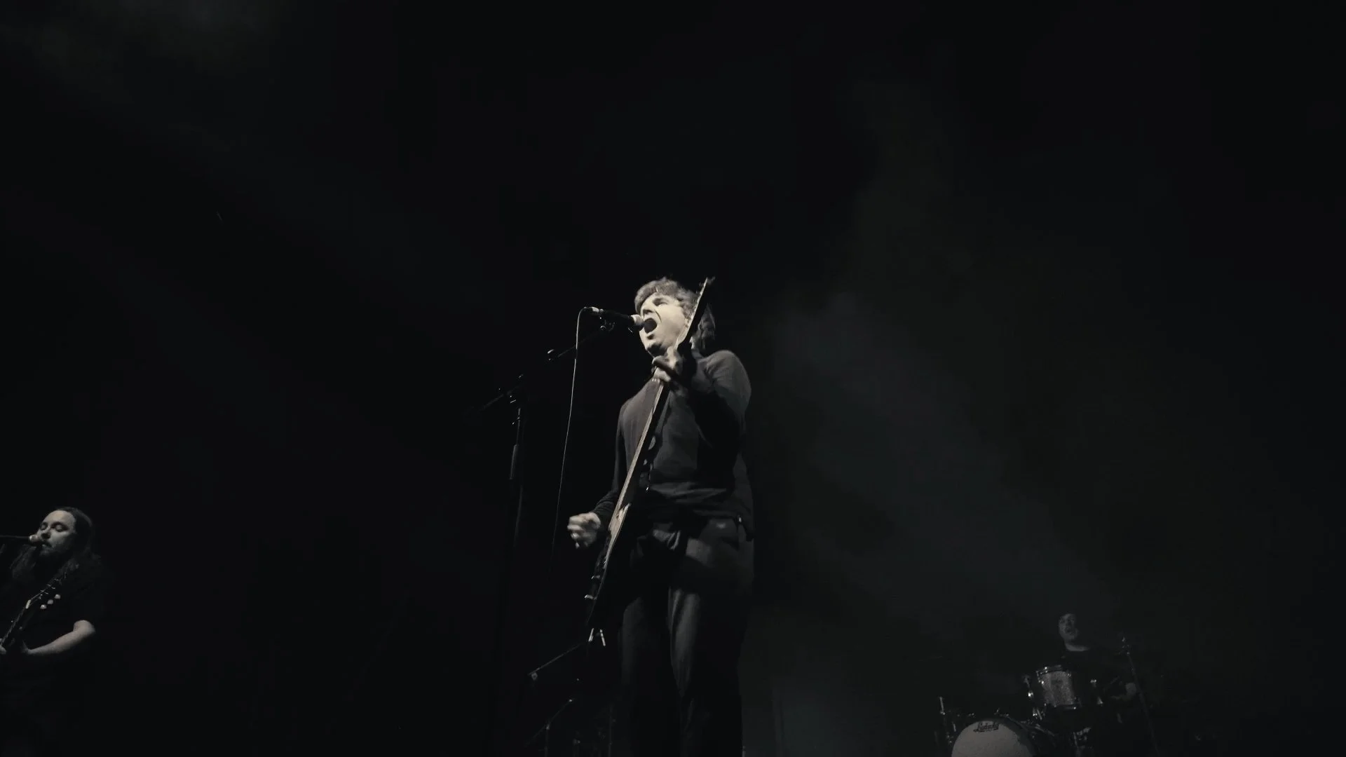 Un groupe de musiciens en performance, un chanteur avec guitare en avant-plan, exprimant de l'émotion, en noir et blanc.