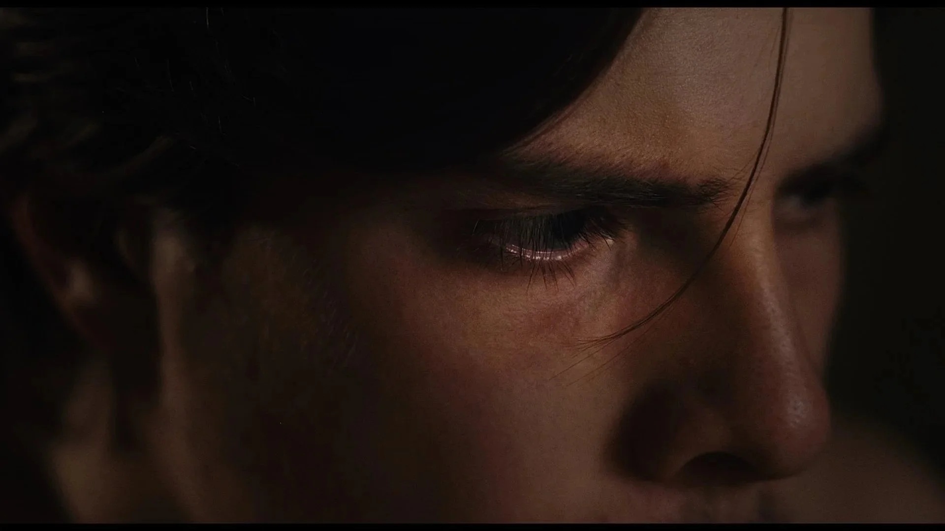 Première plan d'un visage humain, avec un focus sur un œil. La photo est sombre avec un éclairage doux mettant en valeur la peau et les détails de l'œil.