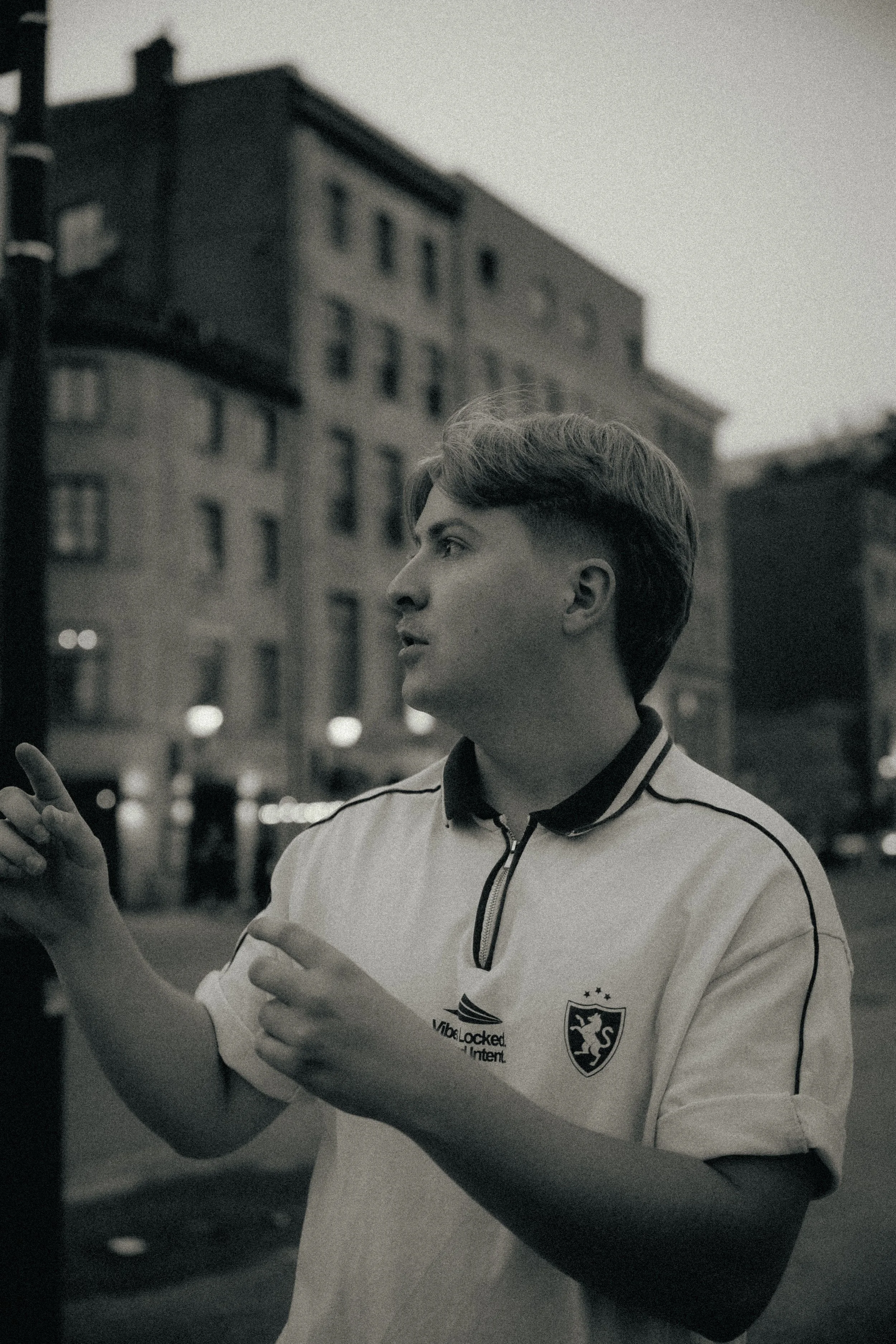 Un jeune homme avec des cheveux courts et une veste de sport blanche, portant un logo d'équipe de football, semble faire un geste ou une communication avec ses mains, dans une scène en noir et blanc en environnement urbain le soir.
