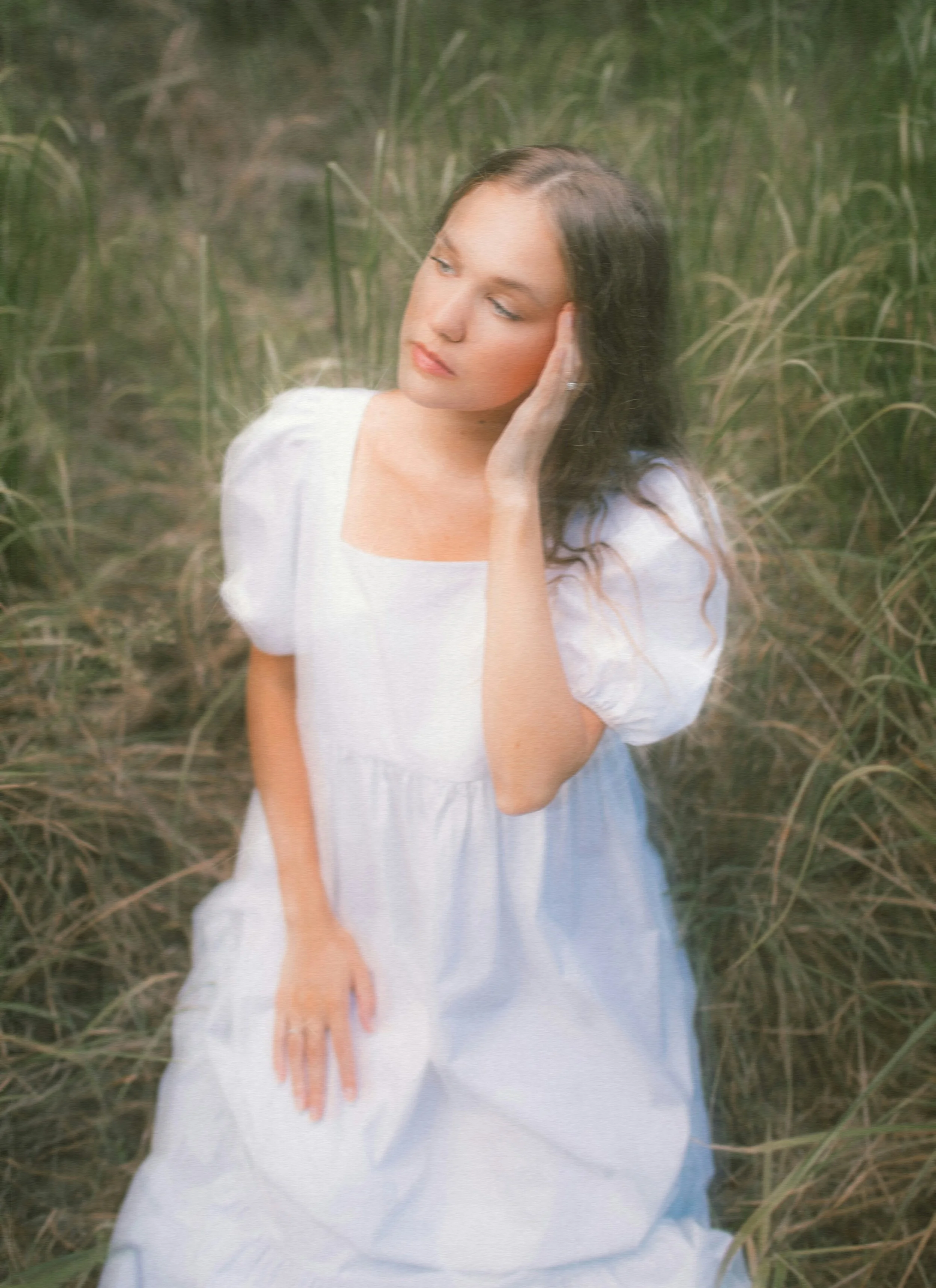A woman with long dark hair, wearing a white dress with puffed sleeves, sitting in a field of tall grass, with her hand gently touching her face and looking contemplative.