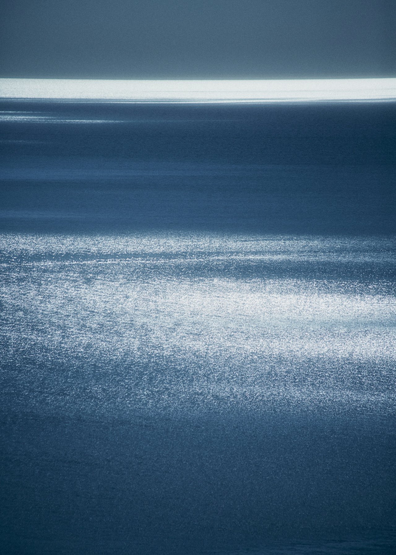 A serene body of water with sunlight reflecting off its surface, creating a shimmering effect, with a clear sky above.