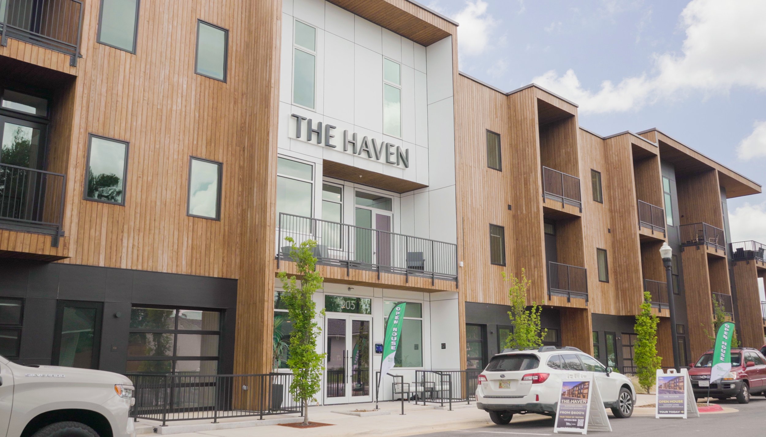 Modern multi-story apartment building with a wood and white exterior, balconies, and large windows, with cars parked in front and signs advertising an open house.