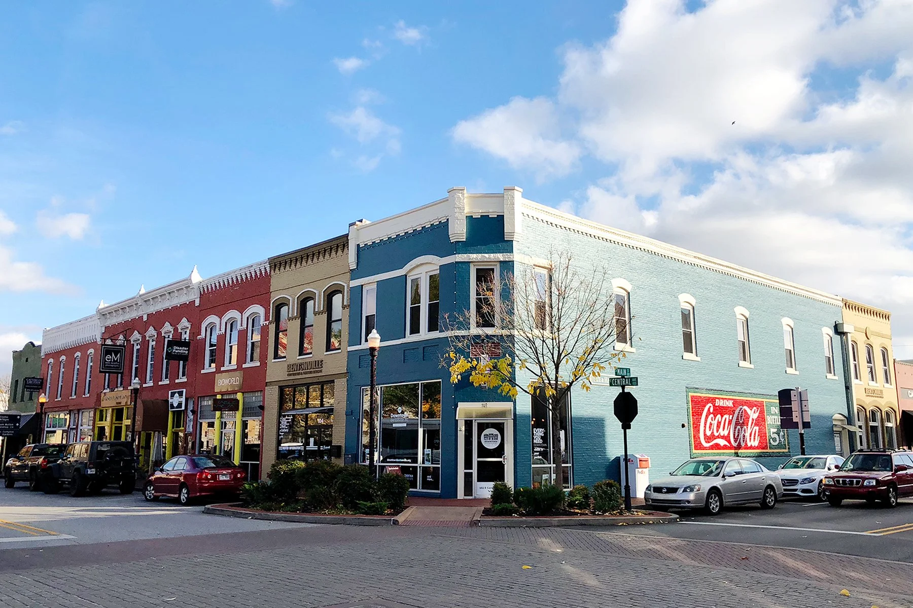 Colorful commercial buildings on a city street with parked cars, a tree with yellow leaves, a Coca-Cola sign, and a street sign at the corner of Main and Central streets, under a partly cloudy sky.