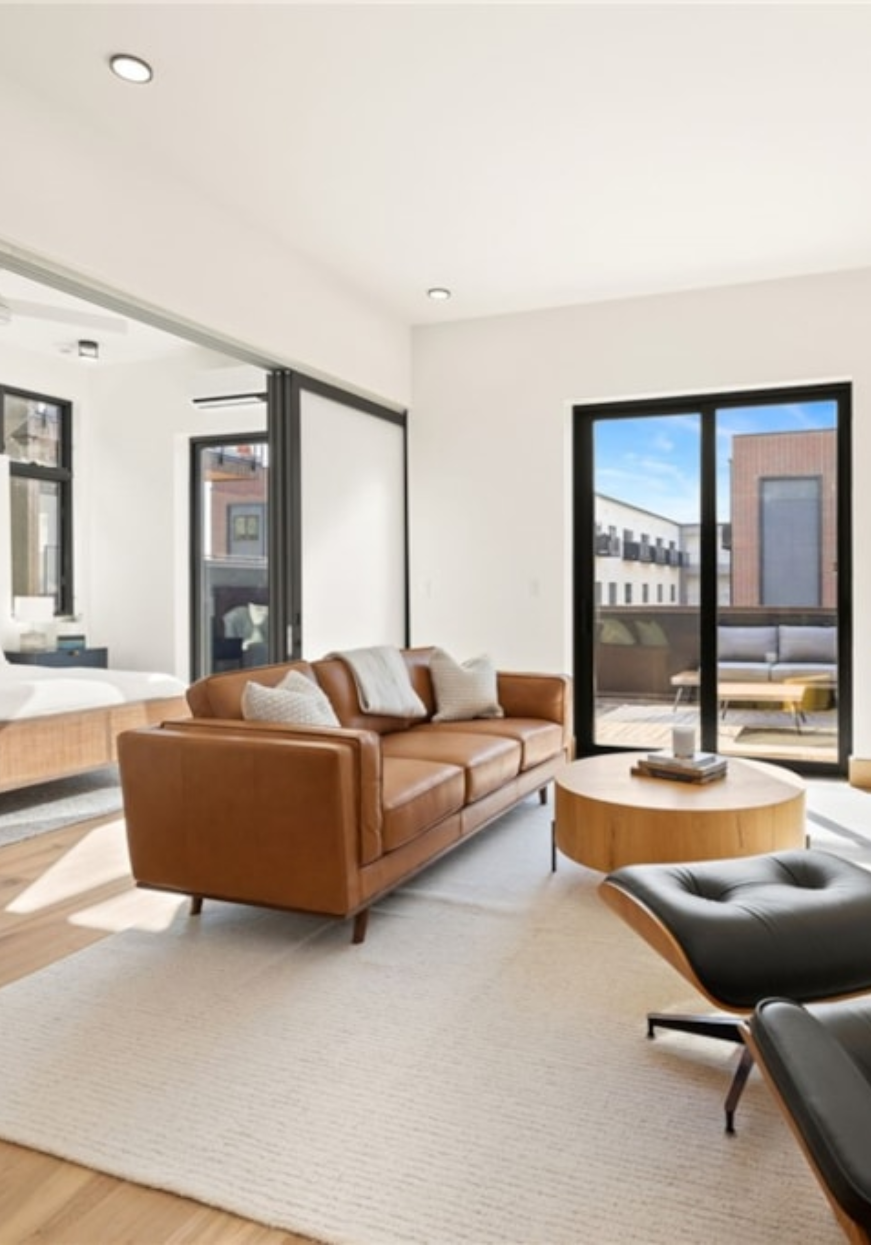 Modern living room with a brown leather sofa, black lounge chair, and large windows showing an urban exterior.
