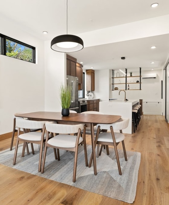 Modern dining area with a wooden table, six white and wood chairs, and a gray area rug. The room features hardwood floors, a black ceiling light, and a window. The open kitchen has wooden cabinets, a white island with a faucet, and open shelves with 