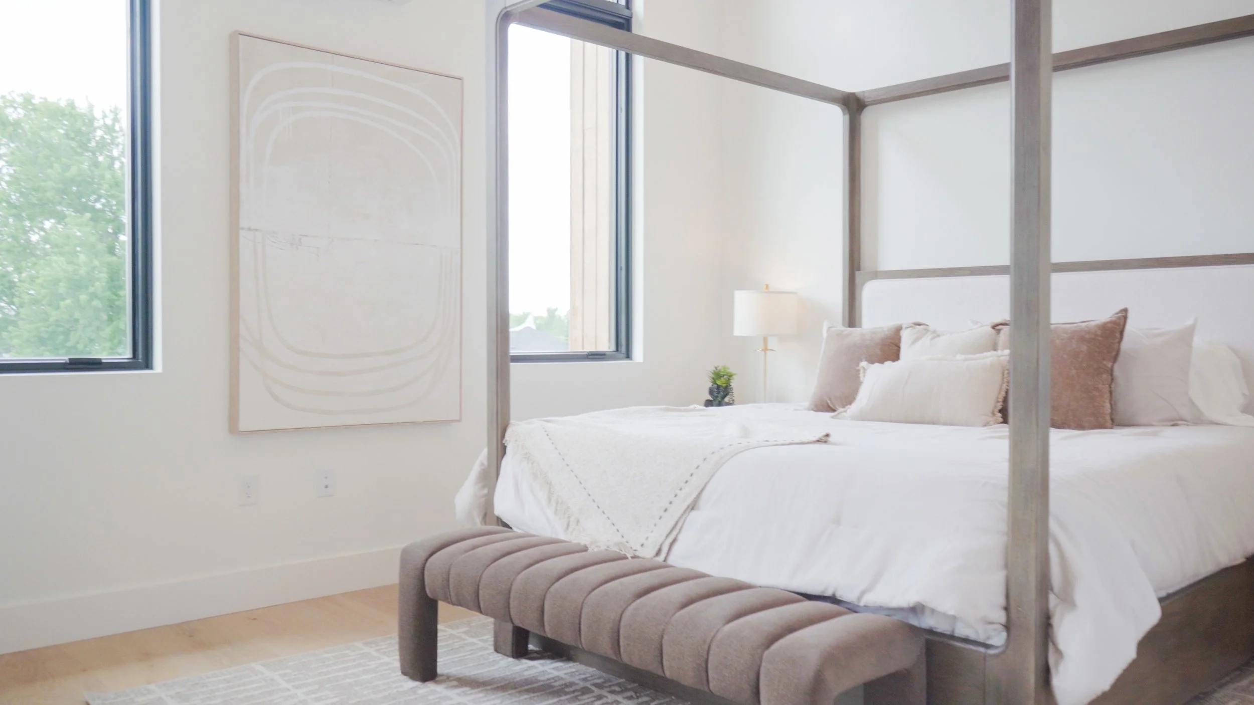 Modern bedroom with large windows, a white bed with neutral pillows, a bedside lamp, a small potted plant, and abstract art on the wall.