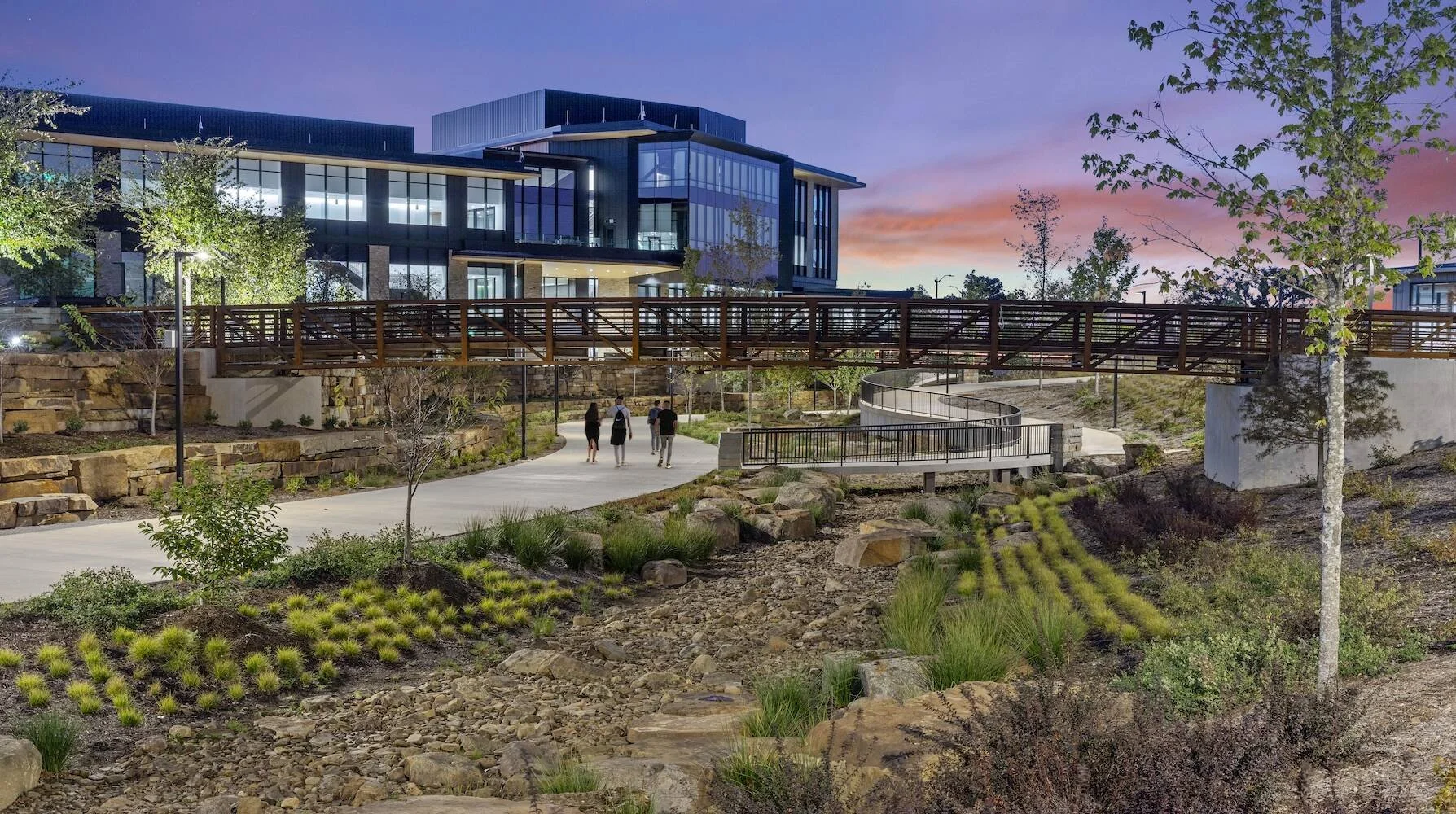 Modern building with large glass windows, a pedestrian bridge, and landscaped park with trees, plants, and a winding stone path, during dusk with colorful sky