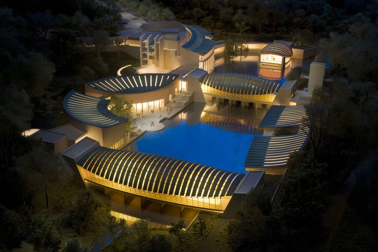 Aerial view of a large, modern building complex with curved, illuminated roofs, a central swimming pool, and surrounding trees at night.