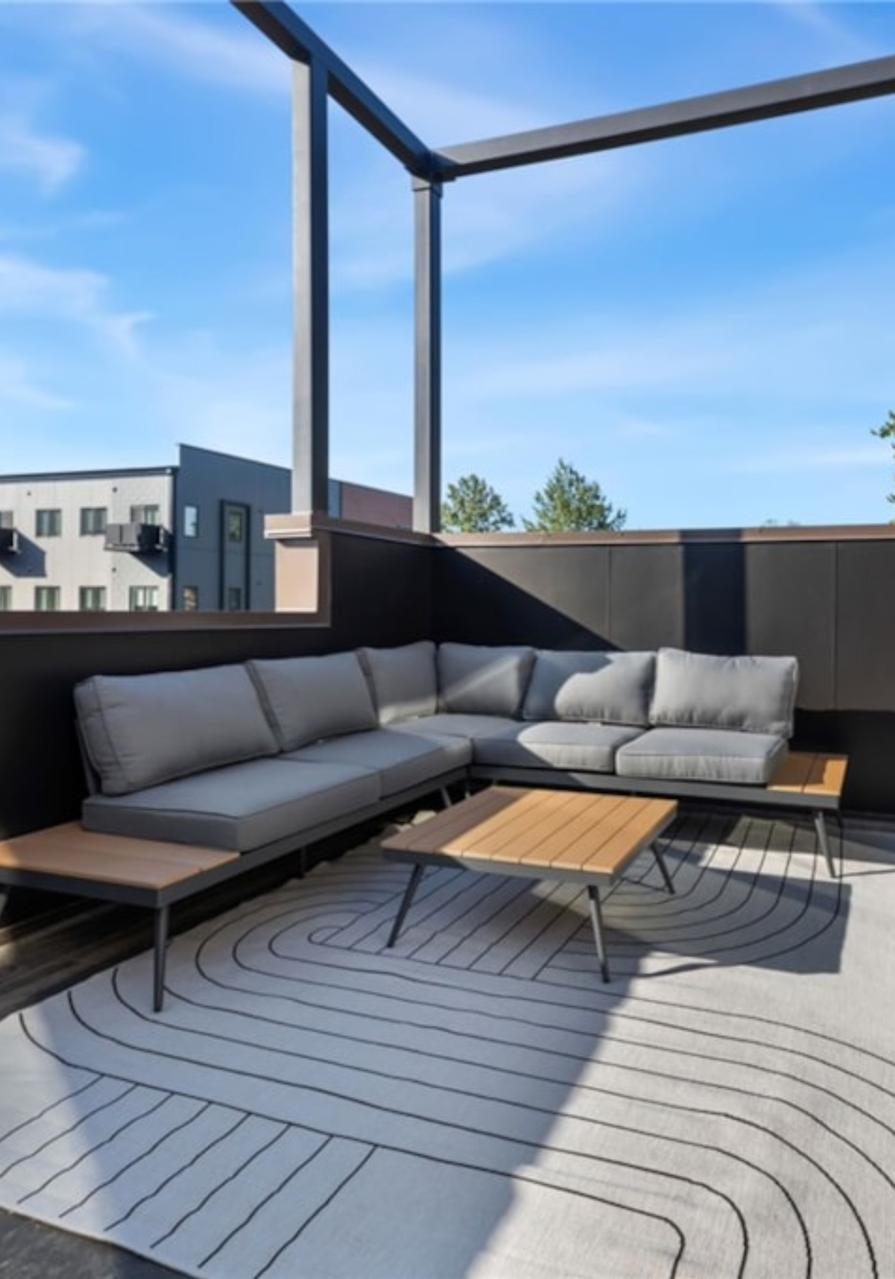 Outdoor rooftop patio with a sectional gray outdoor sofa, two small wooden coffee tables, a black and white patterned rug, and a white fabric umbrella, with a view of a city apartment building and clear blue sky in the background.