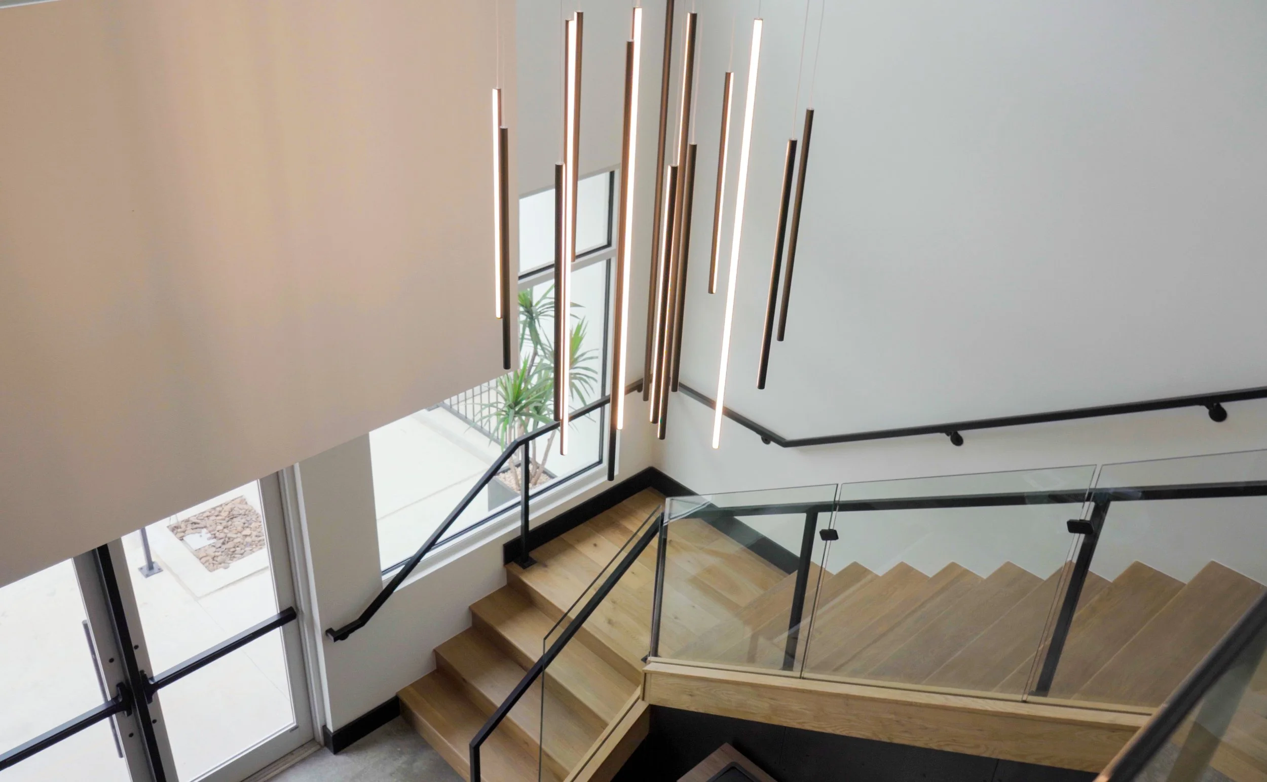 Modern staircase with wooden steps, black handrails, glass panels, and a large ceiling light fixture with multiple hanging cylindrical lights.
