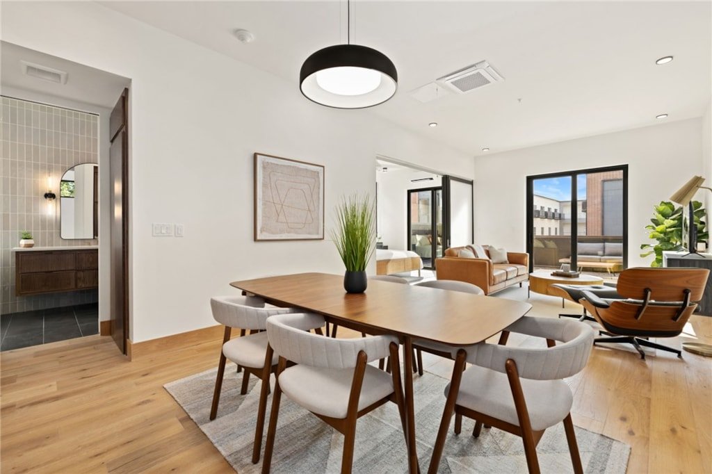 Modern living room with dining area, wooden dining table with white chairs, brown leather sofa, wooden lounge chair, large windows, and sliding glass door, hardwood floors, and decor.